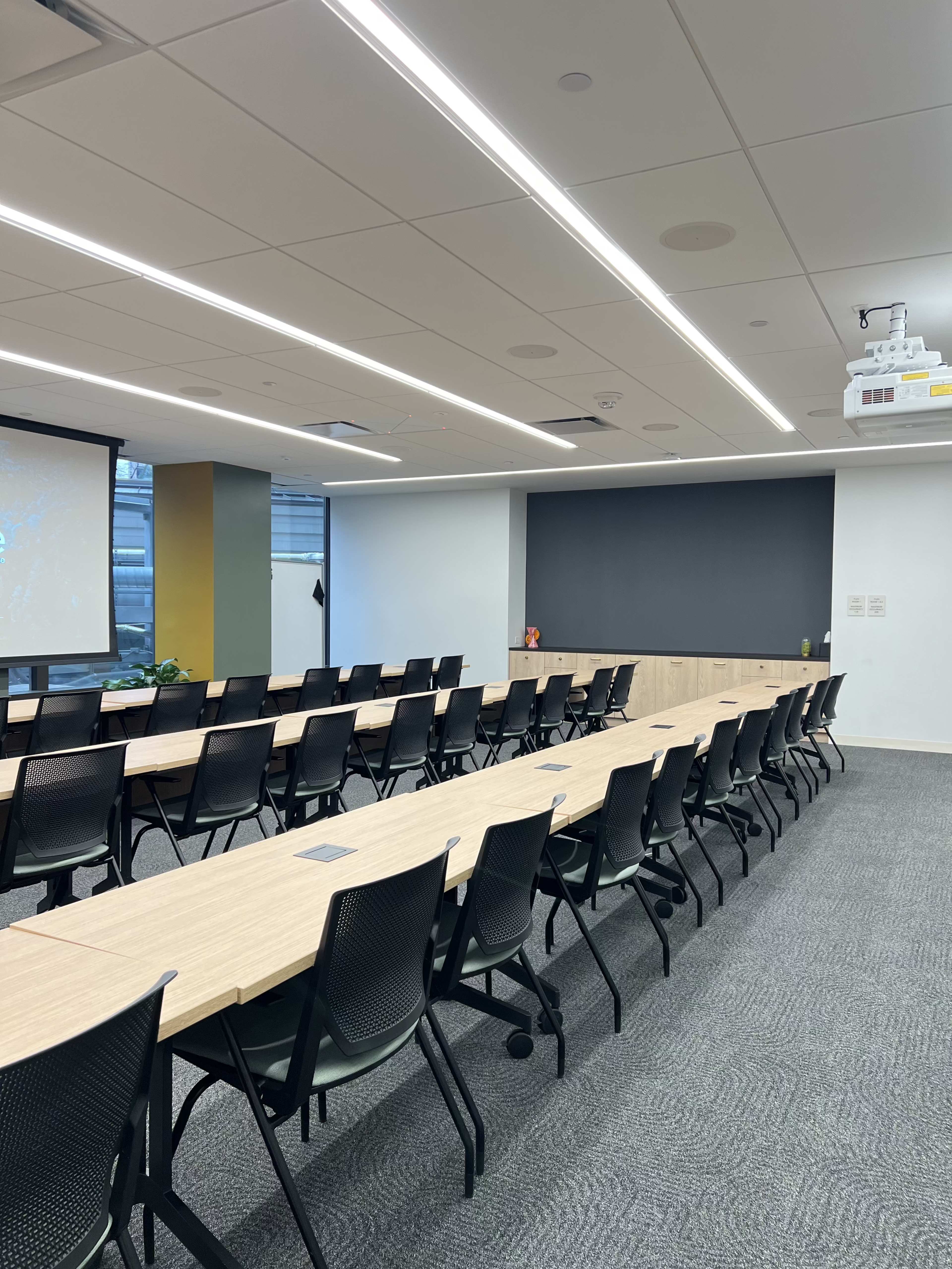 The image shows a modern conference room with multiple rectangular tables arranged in rows and black chairs facing a projector screen.
