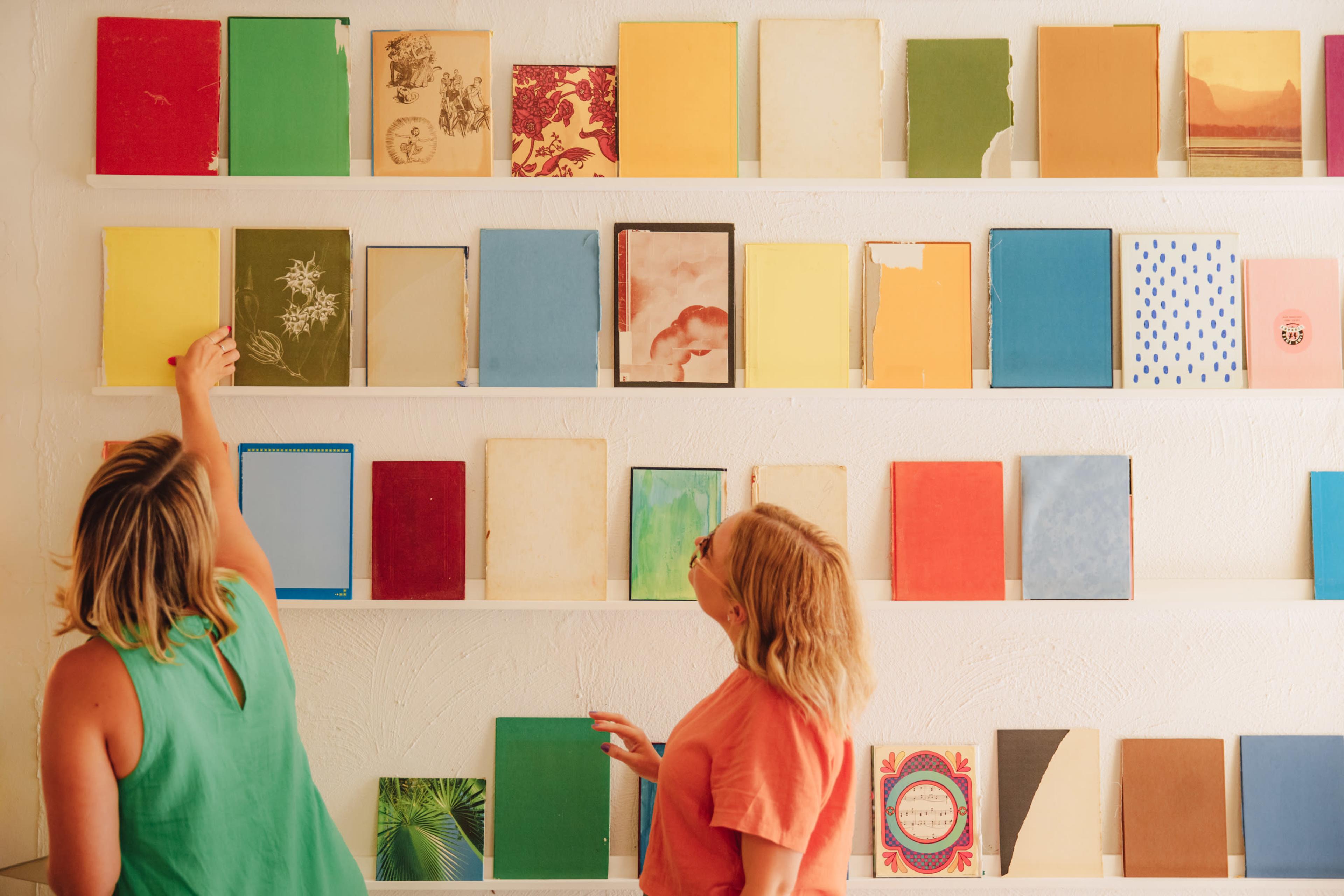 Two women examine colorful books arranged on a white shelf against a textured wall.