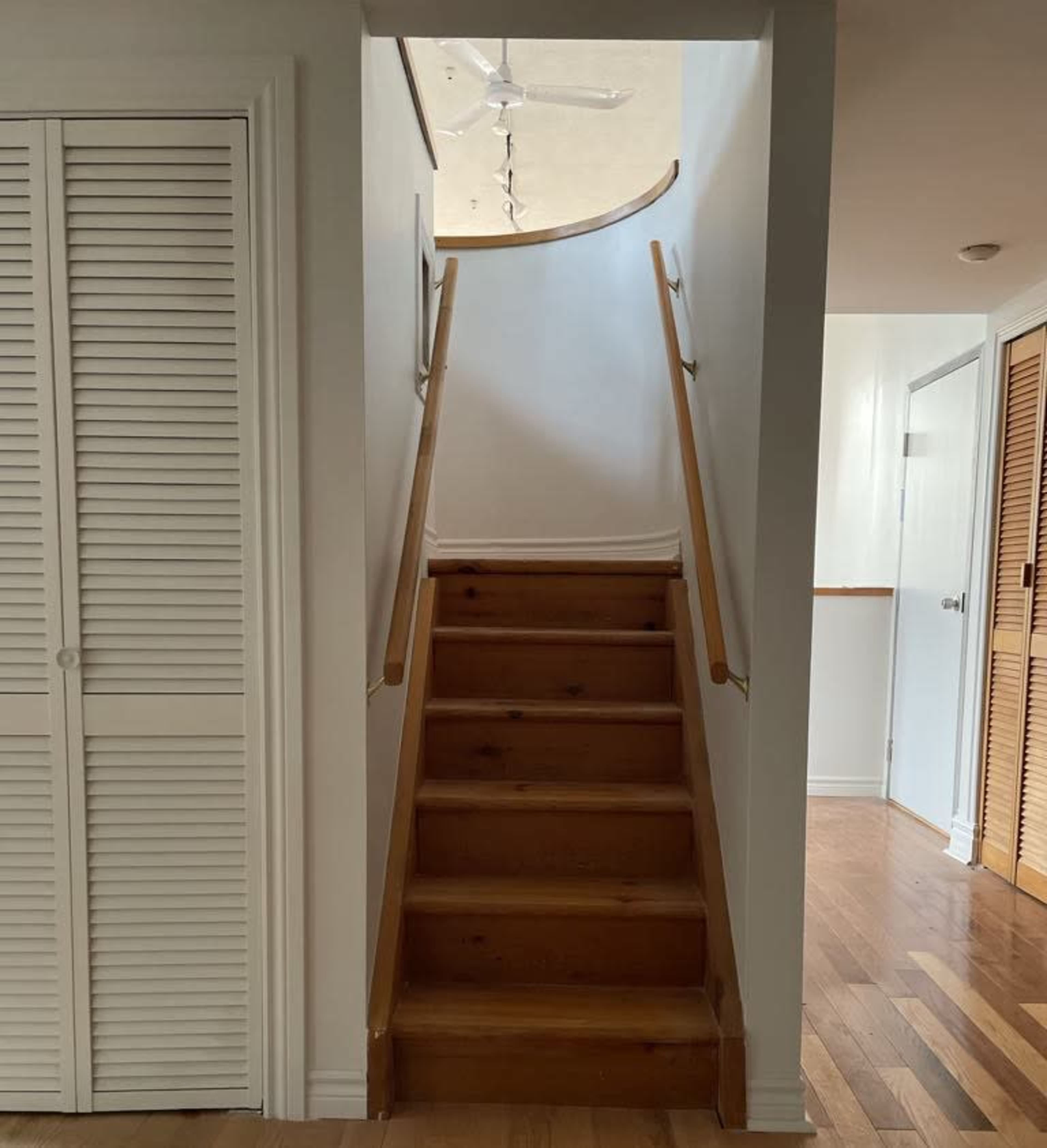 A wooden staircase leads up to a well-lit area with a ceiling fan, framed by simple white walls and doors.