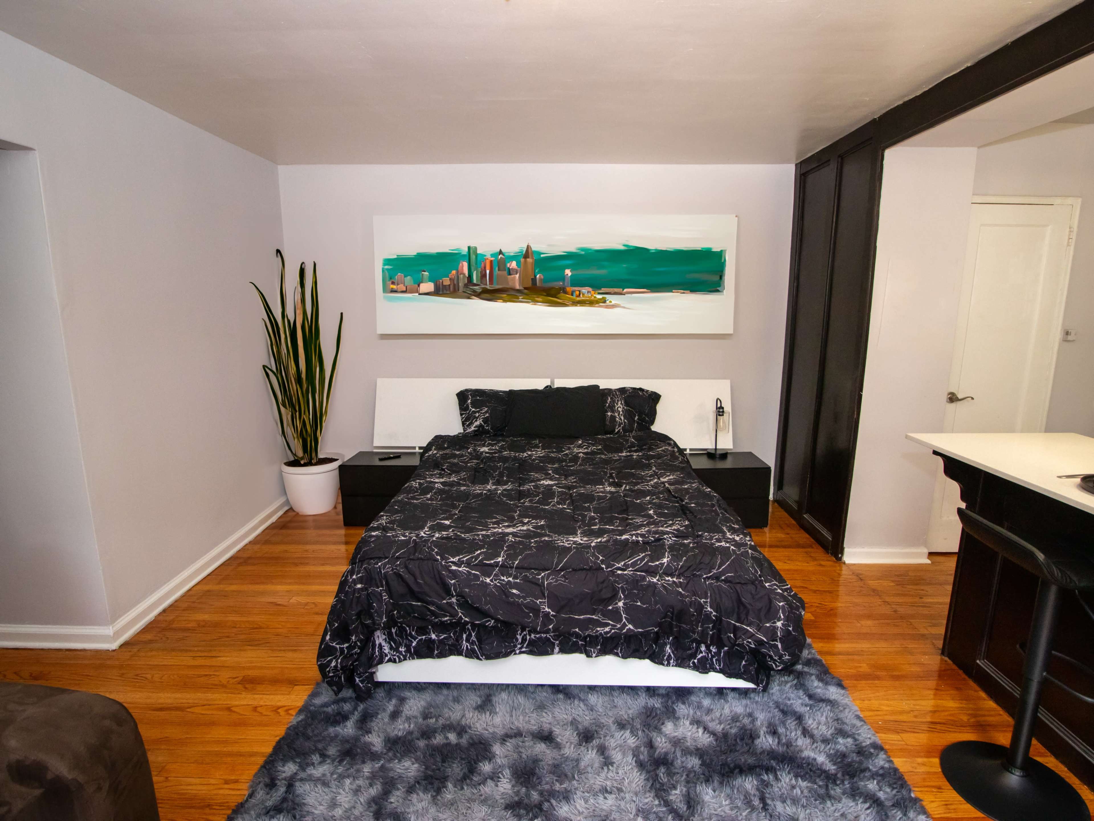 A bedroom features a black and white marble-patterned bedspread, a large painting of a cityscape above the bed, and a potted plant in the corner.