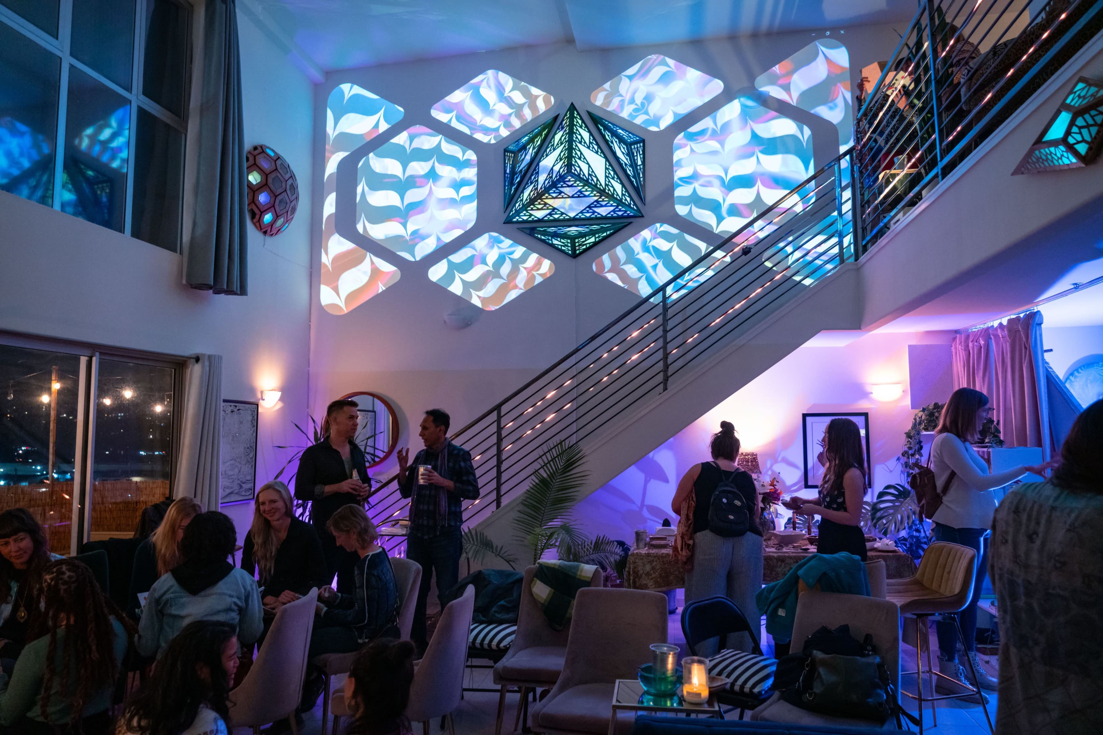 The interior of a modern venue features a staircase adorned with geometric light projections, while guests engage in conversation across various seating areas.