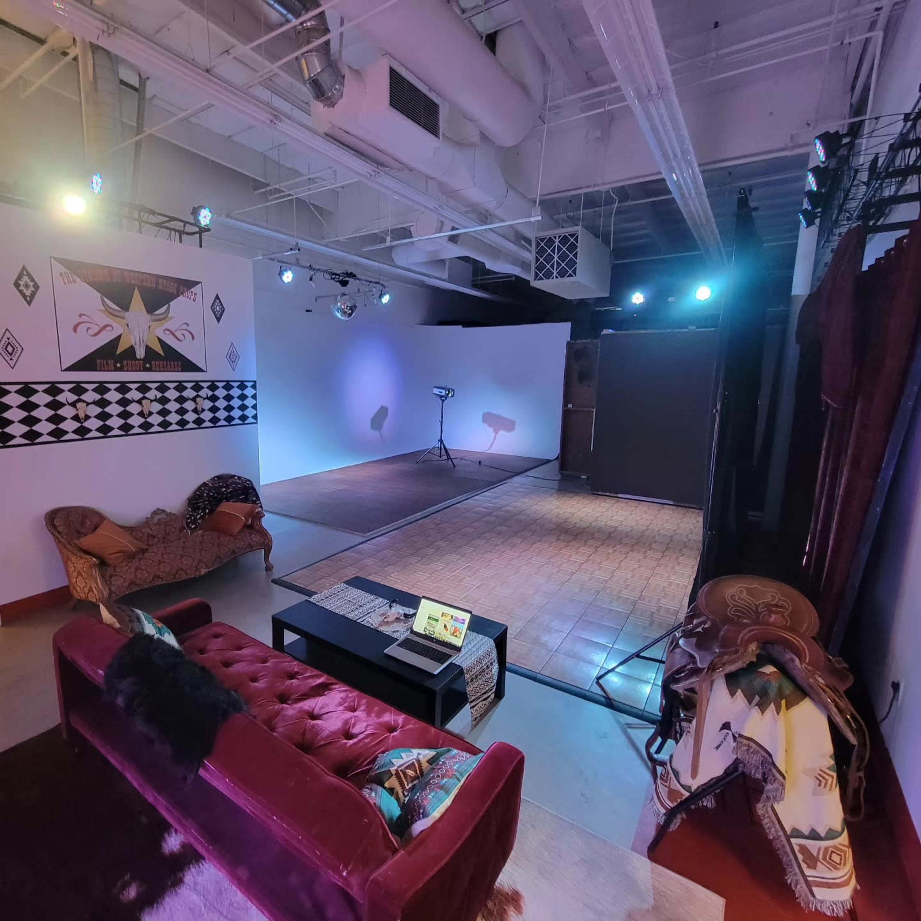 The image shows a modern studio space featuring a red couch, a coffee table with a laptop, and a performance area with stage lighting and a patterned backdrop.