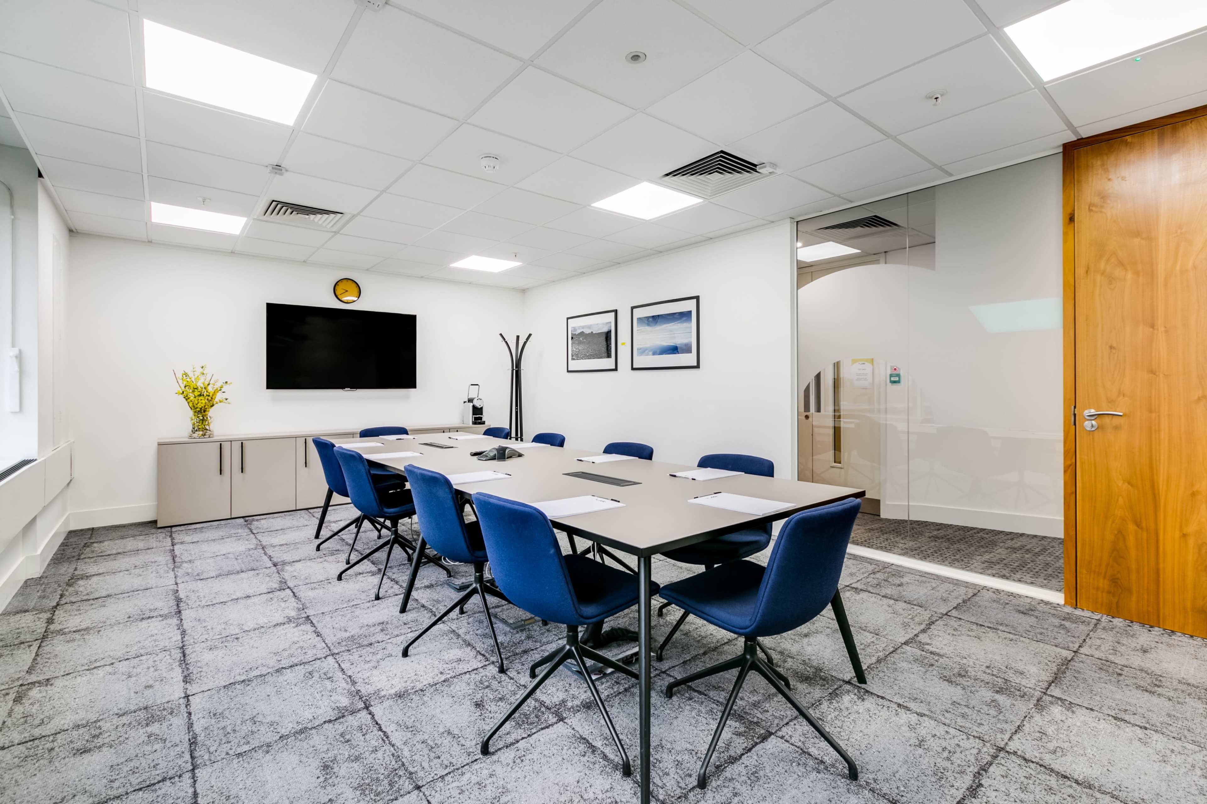 A well-lit conference room features a long table surrounded by blue chairs, a large screen on the wall, and glass partitions.
