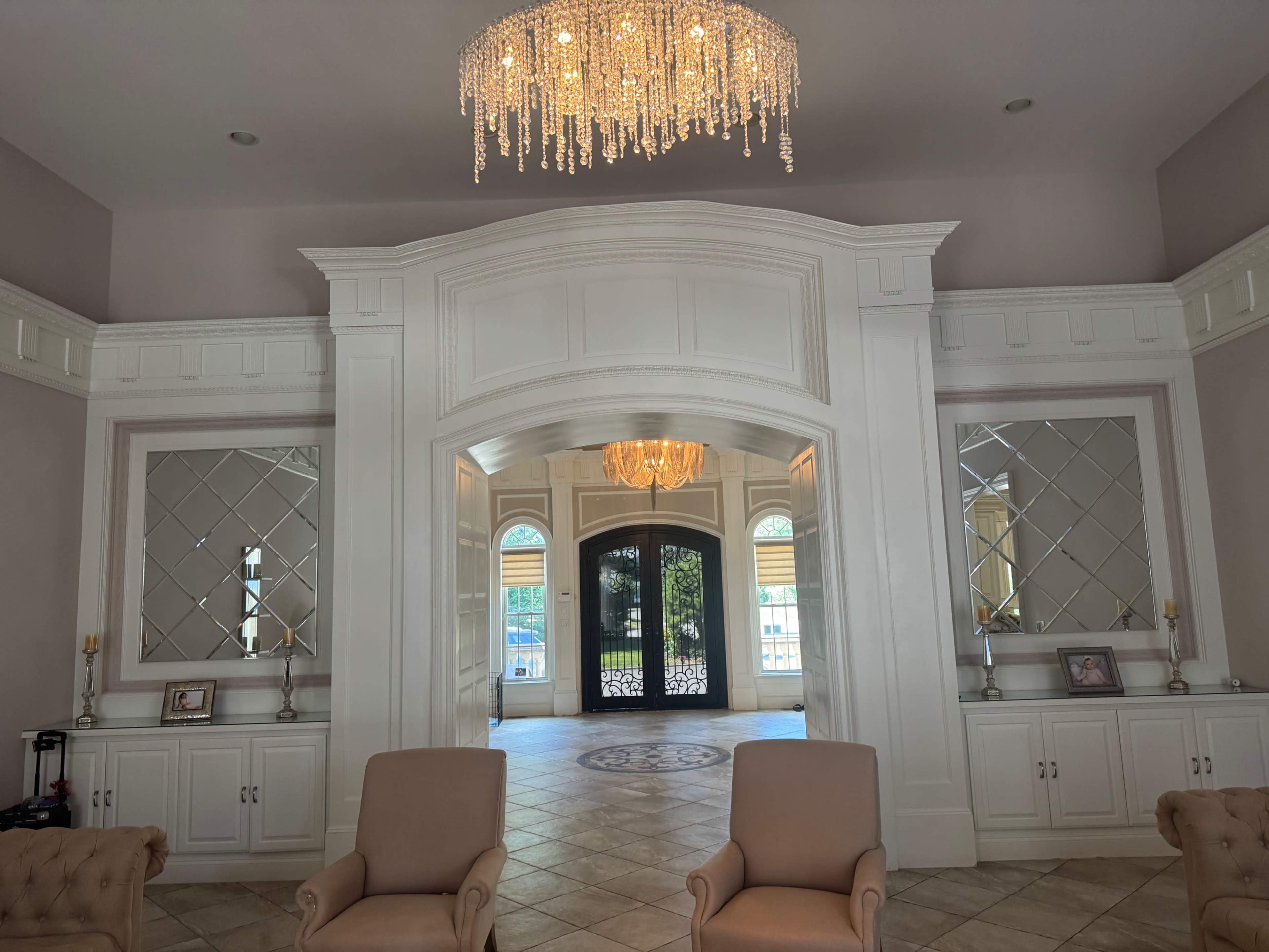 The image shows an elegant foyer featuring two beige armchairs, a crystal chandelier, and mirrored panels on either side of a large doorway.