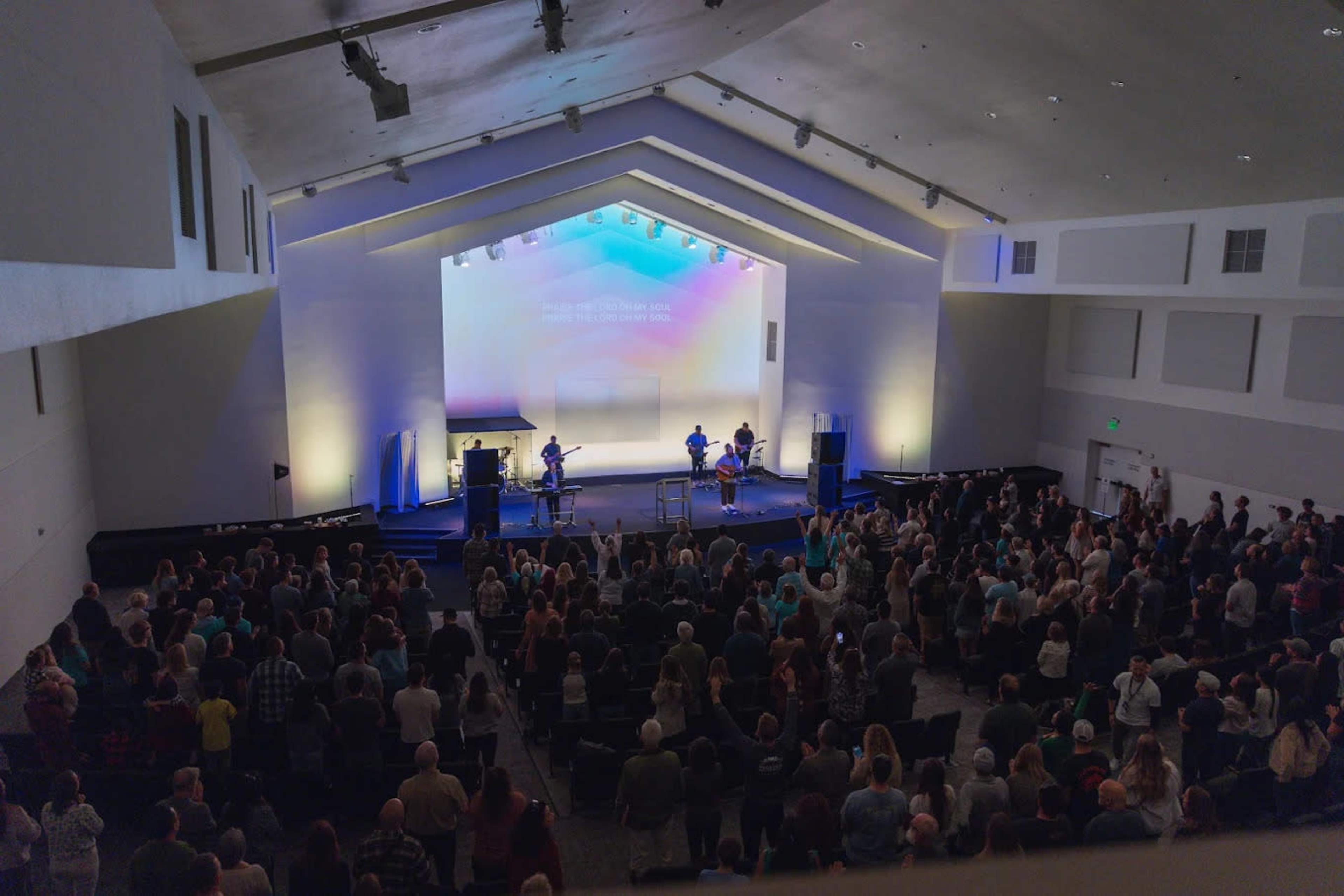 A large congregation gathers in a modern church auditorium as a band performs on stage under colorful lighting.
