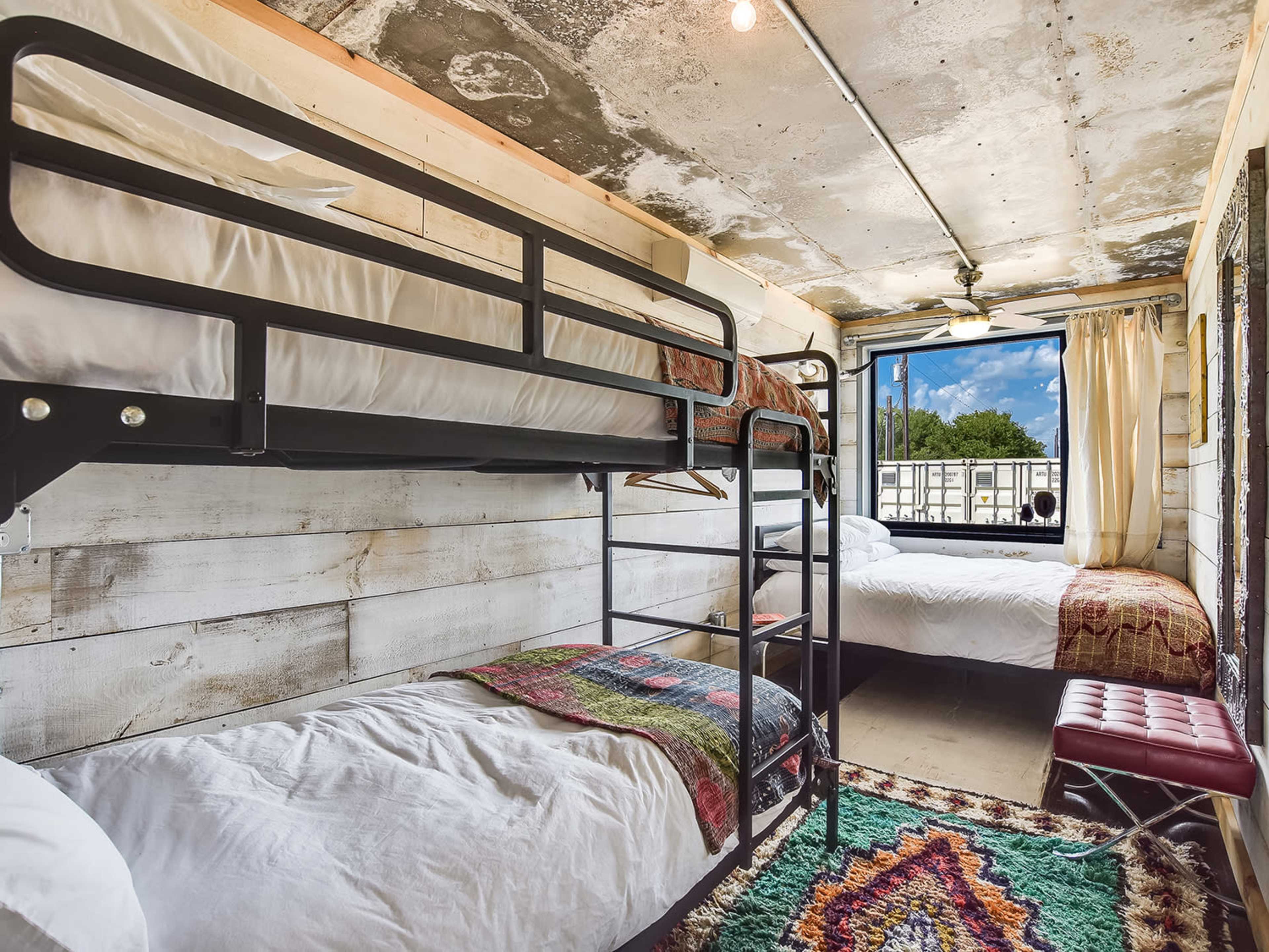 A rustic living space with a bunk bed and a double bed near a window, surrounded by wooden walls and a colorful rug on the floor.