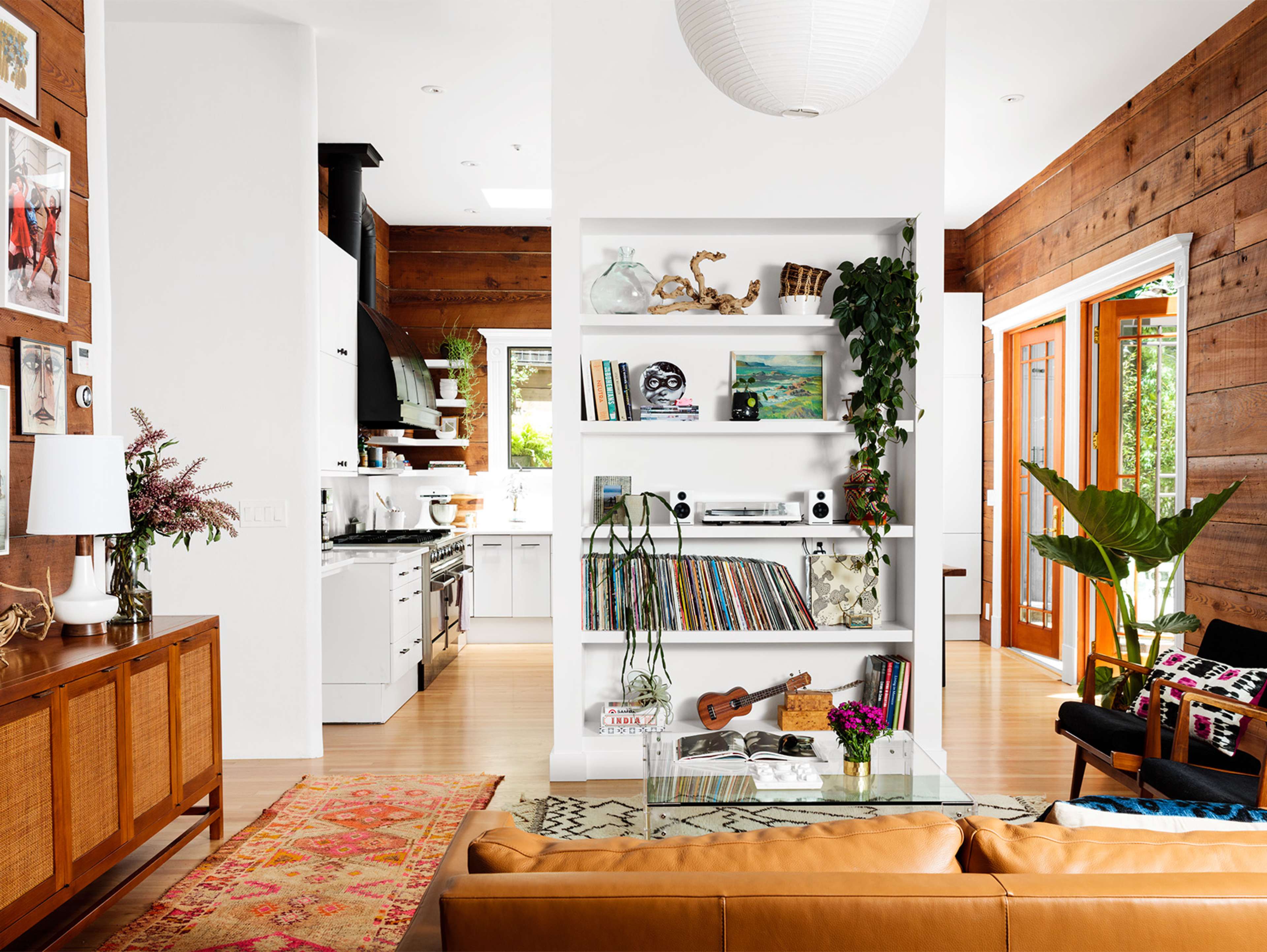 A bright, modern living space with a white shelf filled with books and decor, a cozy brown leather sofa, and an open kitchen in the background.