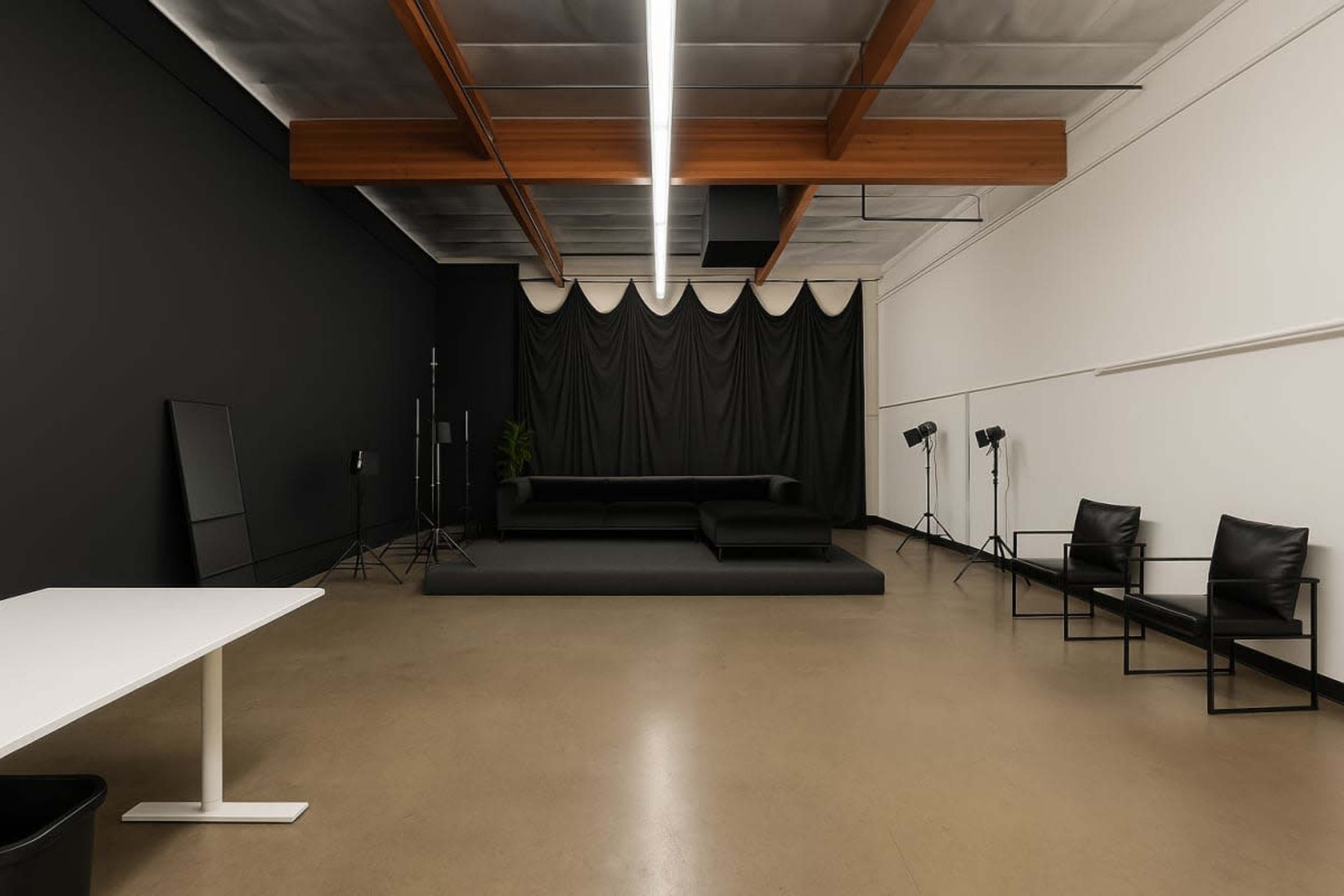 The image shows a spacious studio with a black wall, a large black backdrop, a seating area, and lighting equipment positioned on either side.