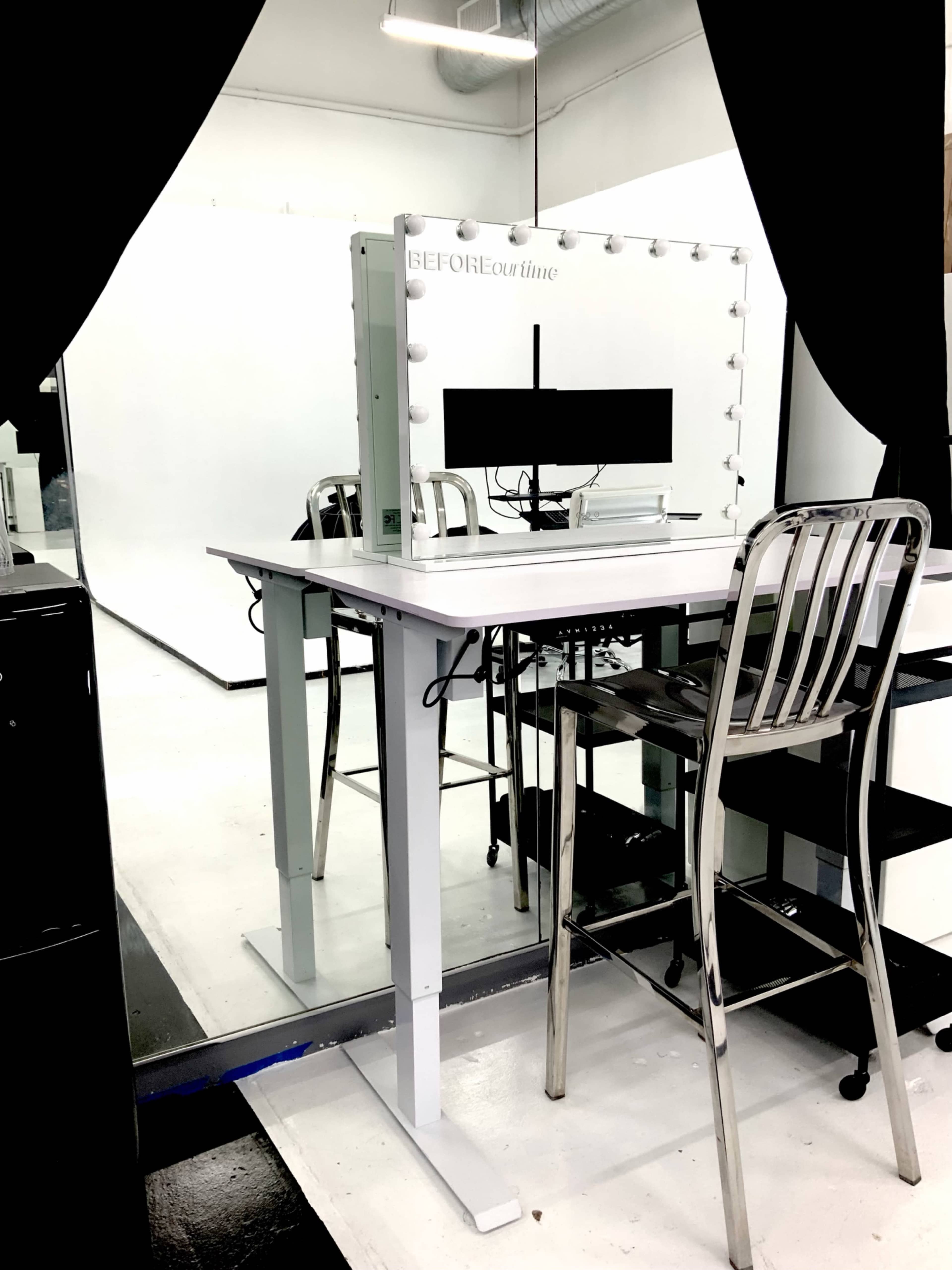 A makeup station with a backlit mirror, two stools, and a monitor is set up in a brightly lit studio space.