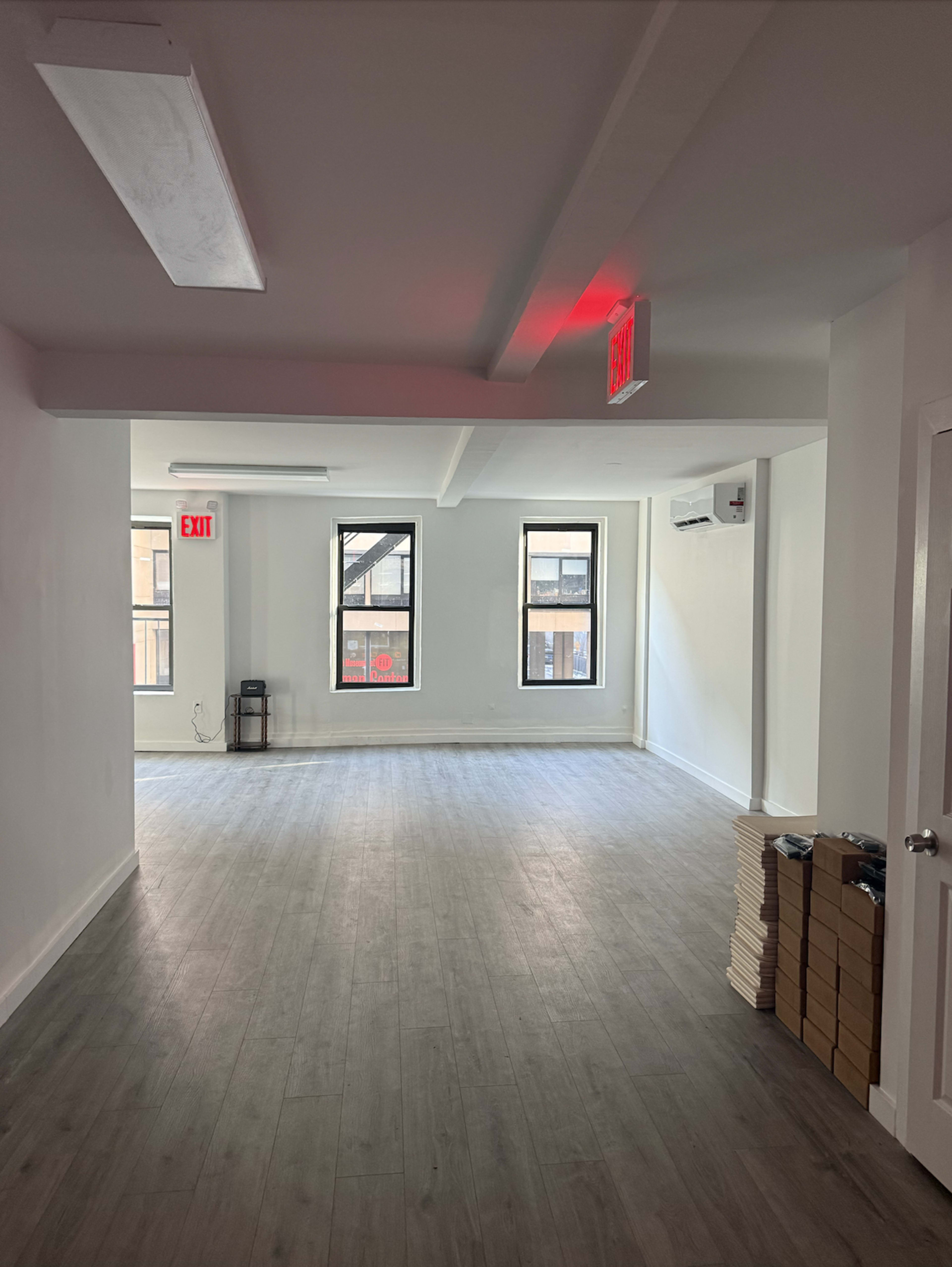 The image shows an empty room with large windows on one side, light-colored walls, and a wood-like floor.