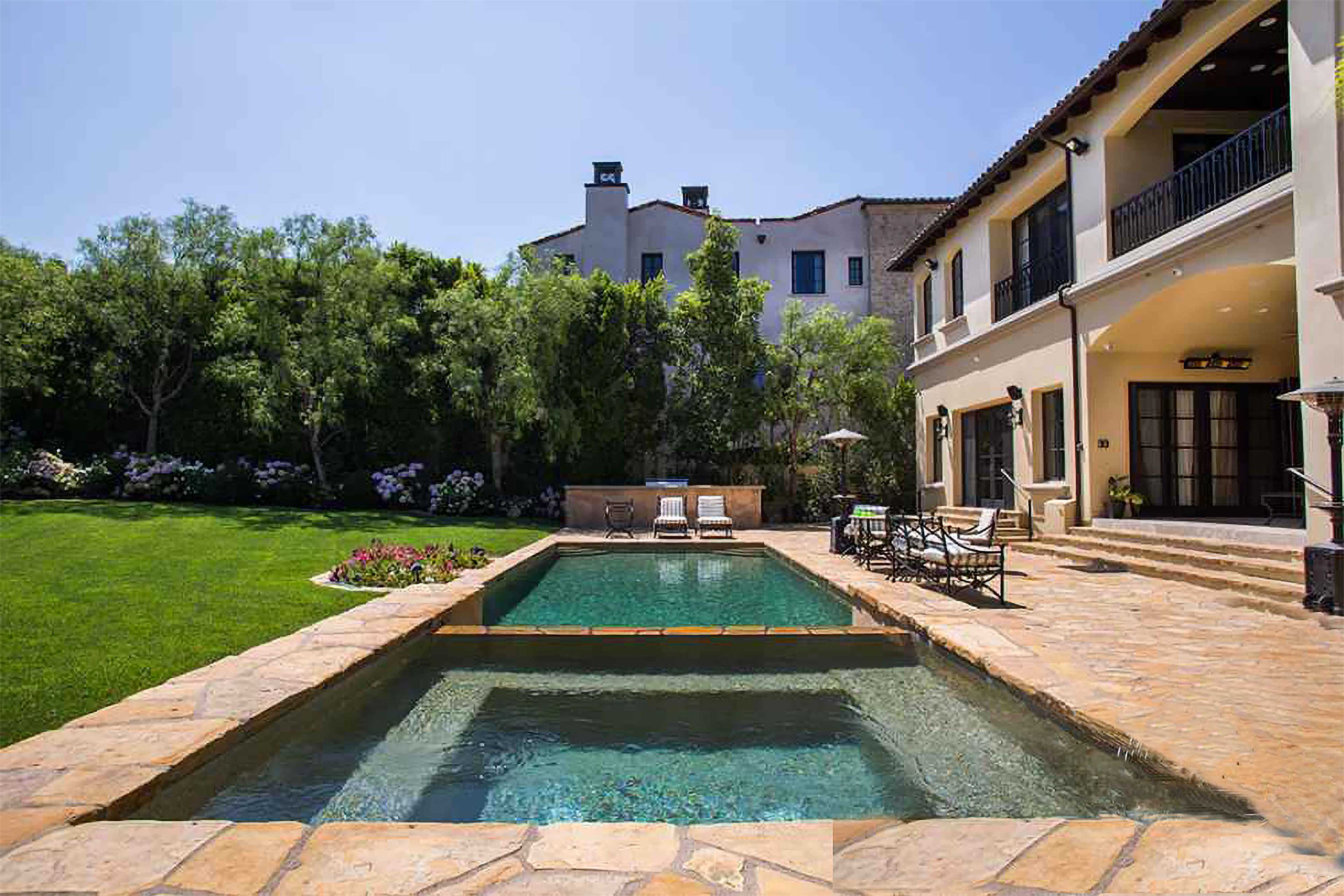 A residential backyard featuring a swimming pool, stone patio, and a well-maintained lawn surrounded by trees and shrubs.