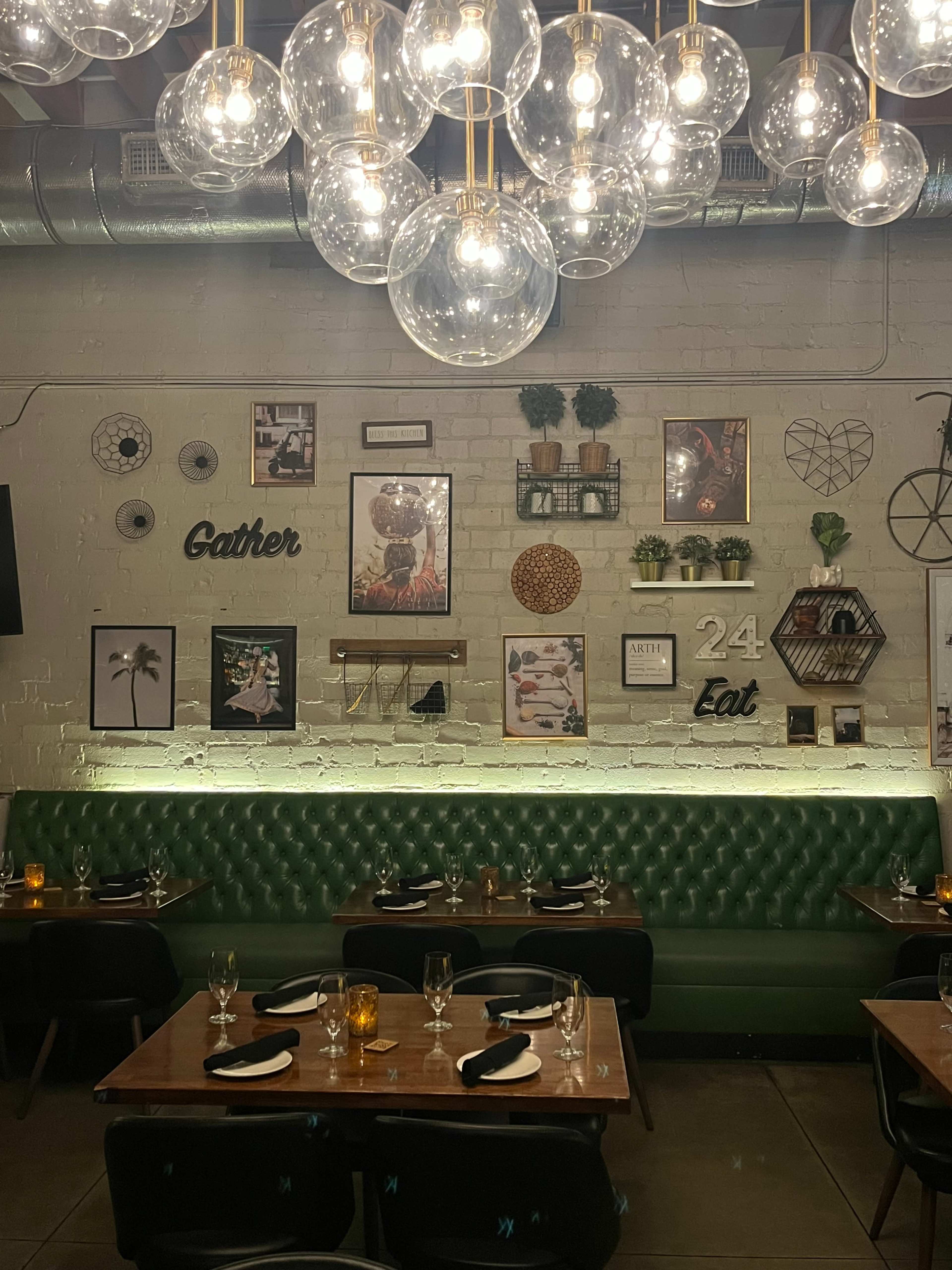 The image shows a restaurant interior featuring green tufted seating, wooden tables set with cutlery, and a wall adorned with various decorative frames and plants.