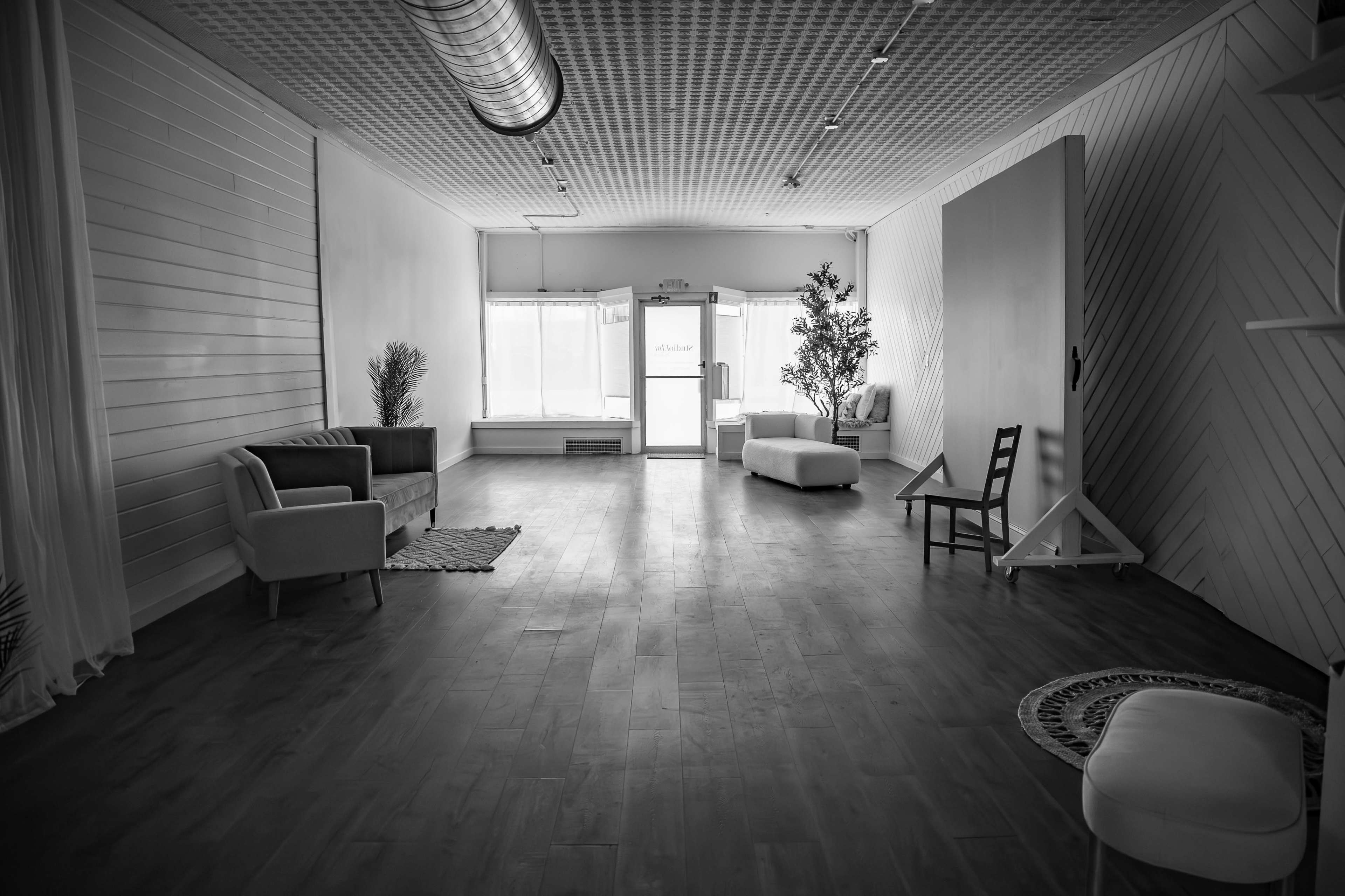 The image shows a spacious, empty room with light-colored wooden flooring, a few simple pieces of furniture, and large windows letting in natural light.