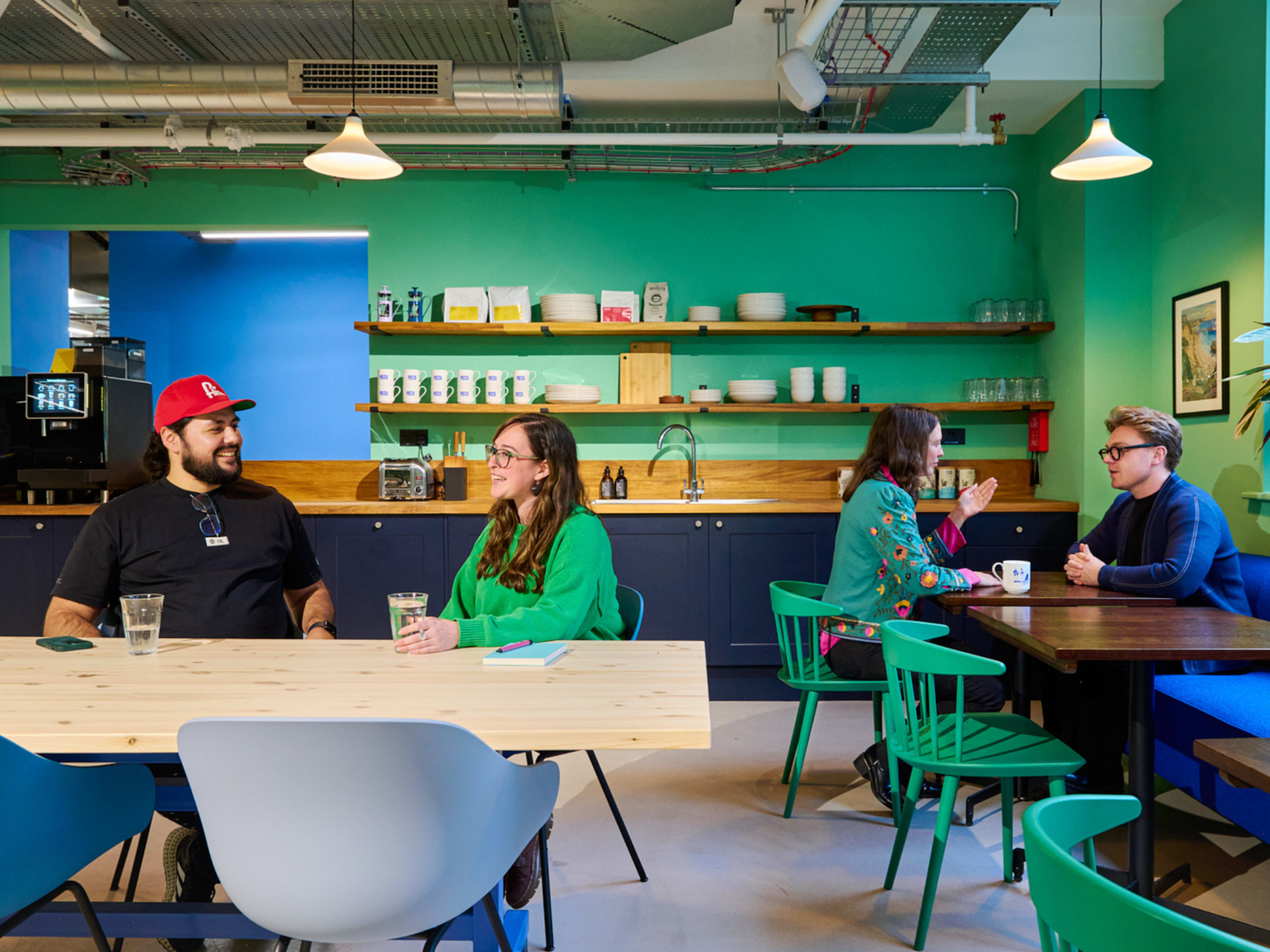 Two groups of people are sitting at tables in a brightly colored communal kitchen, engaged in conversation, while a coffee machine and various kitchen items are visible in the background.