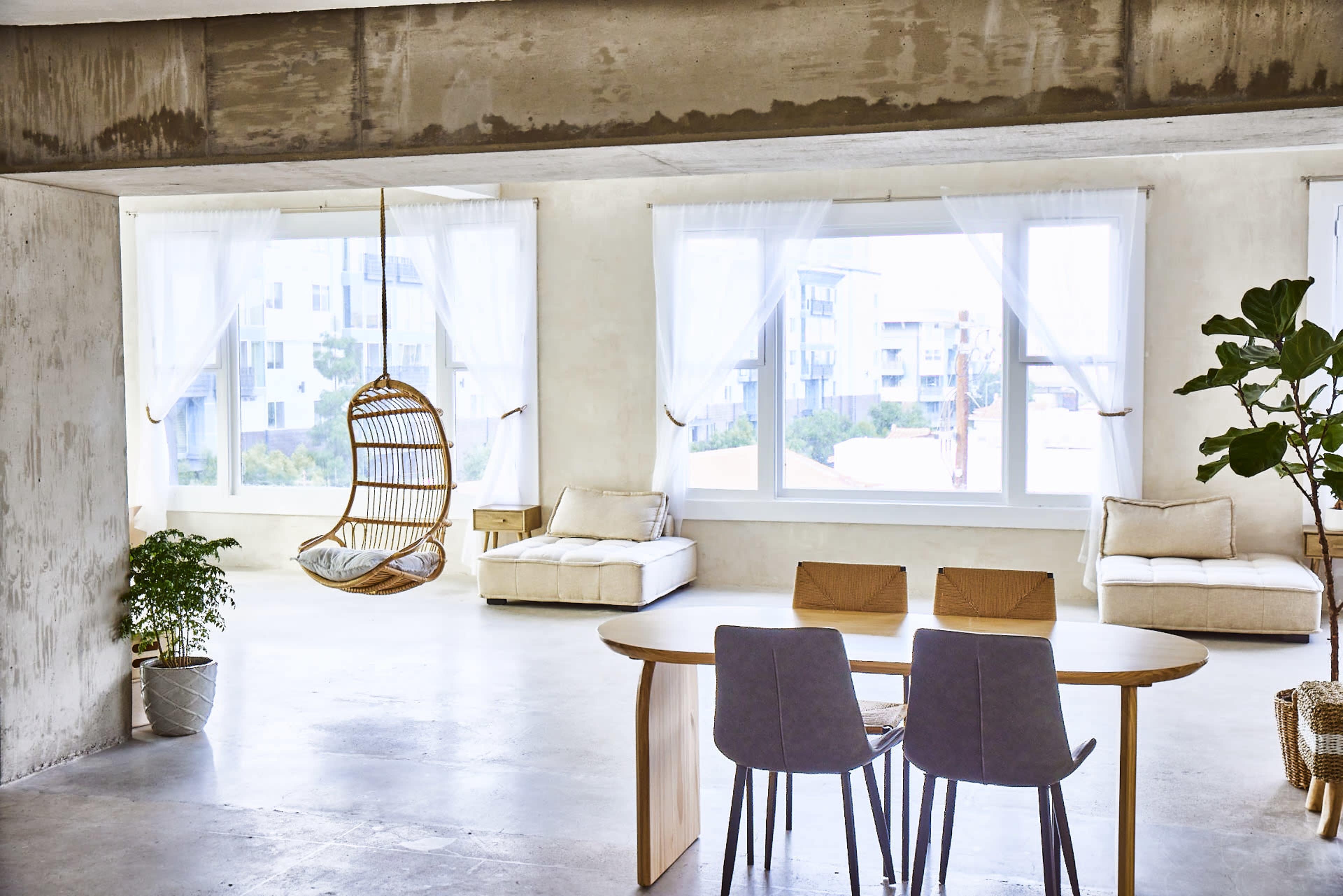 The image shows a minimalist indoor space with a hanging chair, a round dining table with four chairs, and large windows allowing natural light to fill the room.