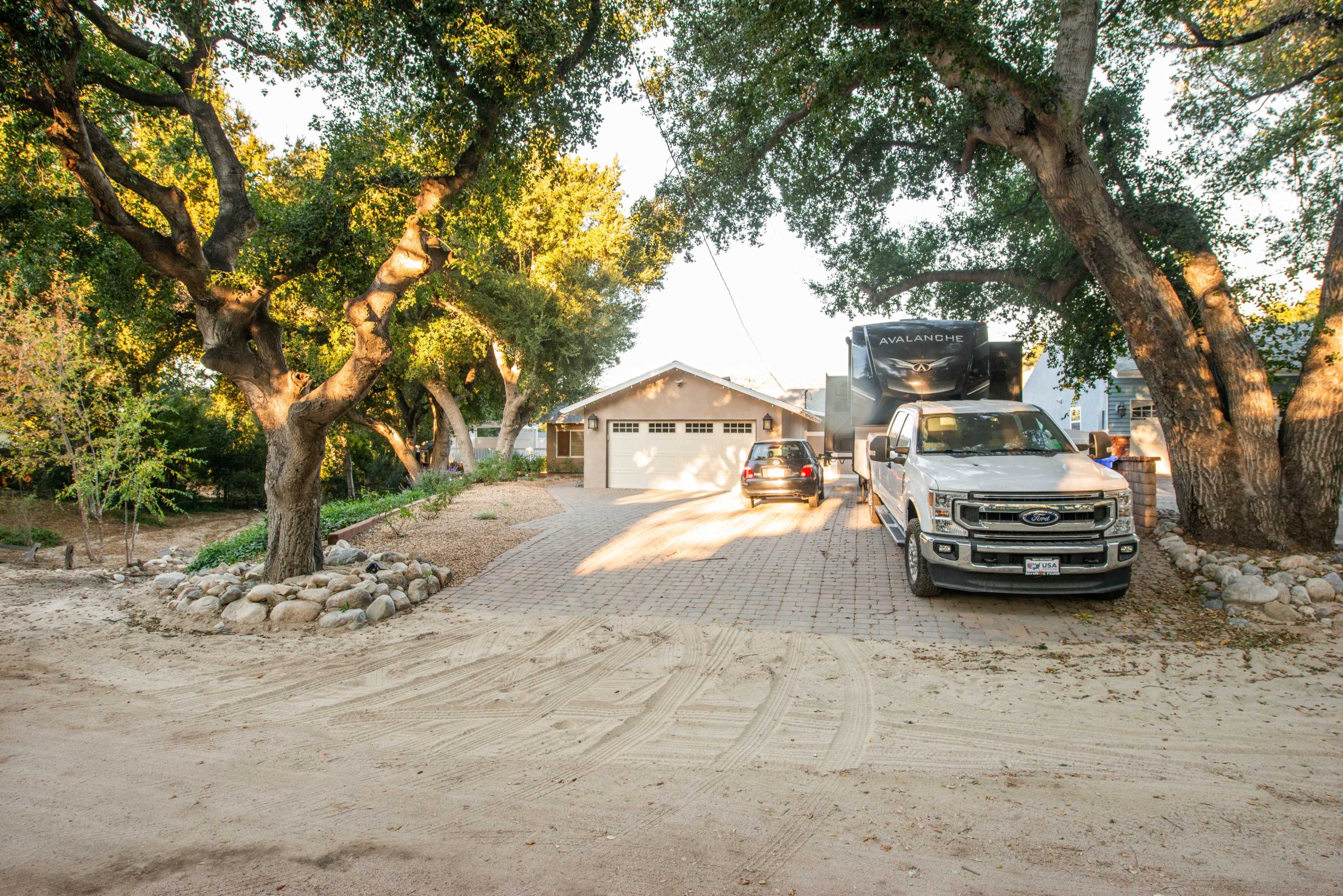 A gravel driveway leads to a house surrounded by trees, with two parked vehicles, including a truck and an RV.