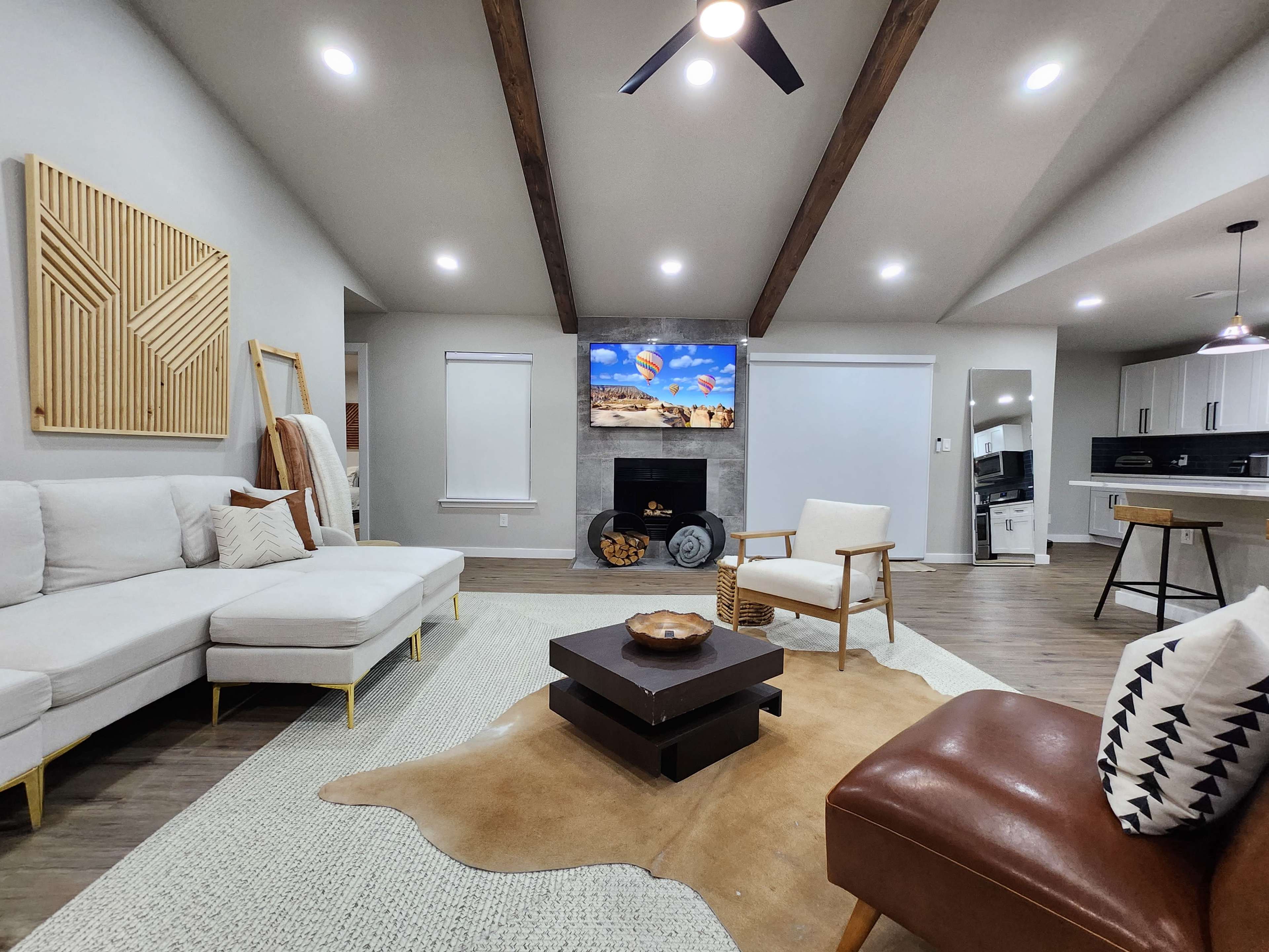 A modern living room with a white sectional sofa, a wooden coffee table, and a wall-mounted television displaying a landscape, all set against a backdrop of exposed wooden beams and light-colored walls.
