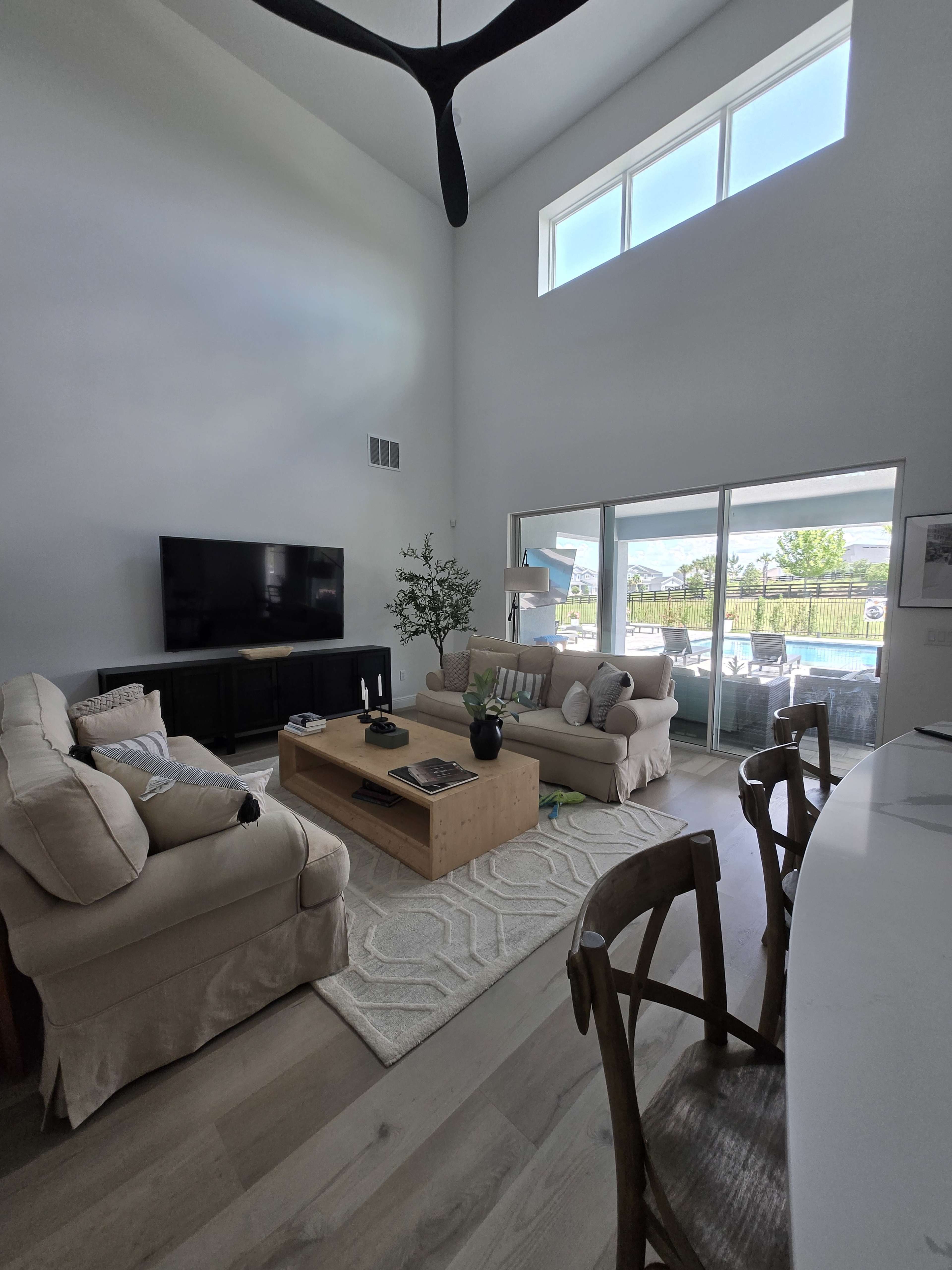 The image shows a modern living room with a large window, a beige sofa and chairs, a wooden coffee table, and a television mounted on the wall.