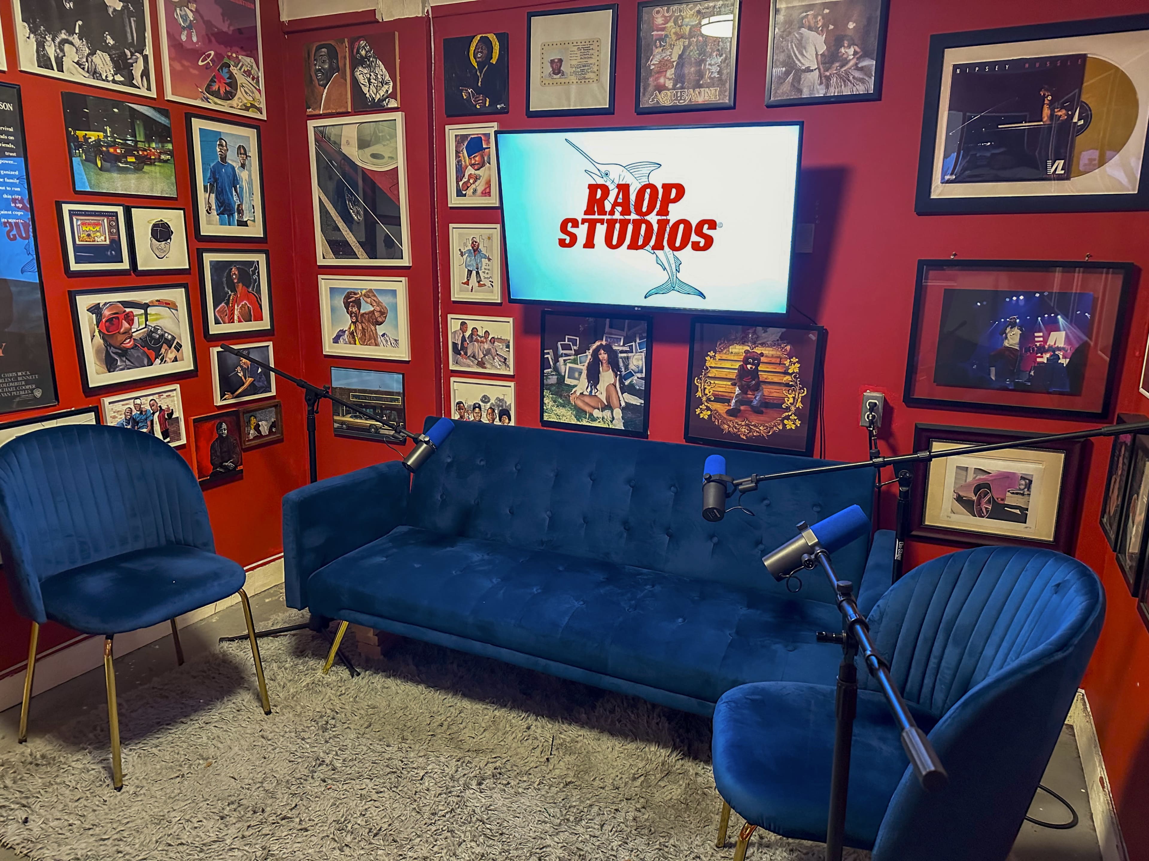 A recording studio decorated with framed photographs and album covers, featuring a blue couch and two matching chairs, along with microphones positioned for podcasting.
