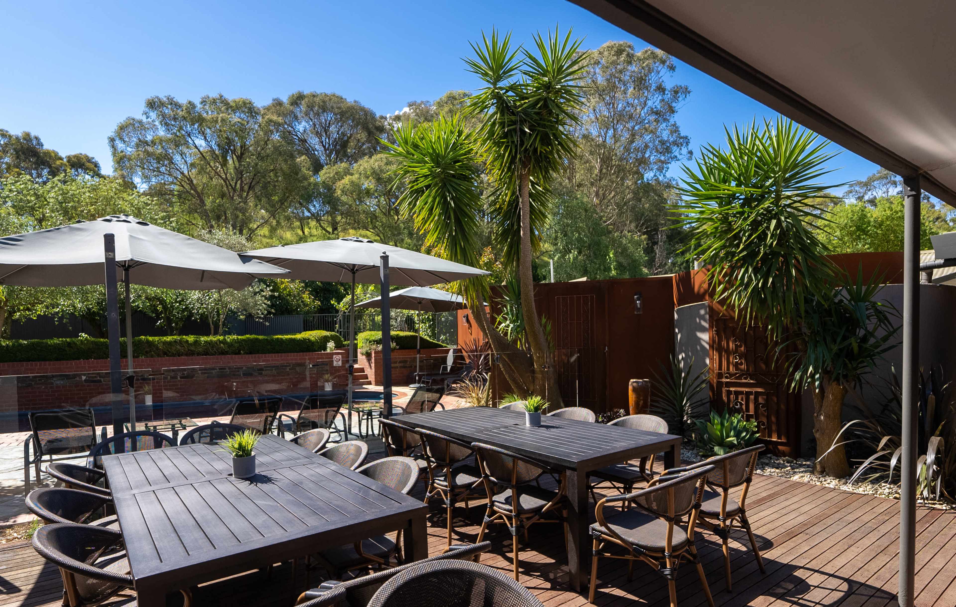 The image shows a patio area with several tables and chairs under large umbrellas, surrounded by greenery and a wooden fence.