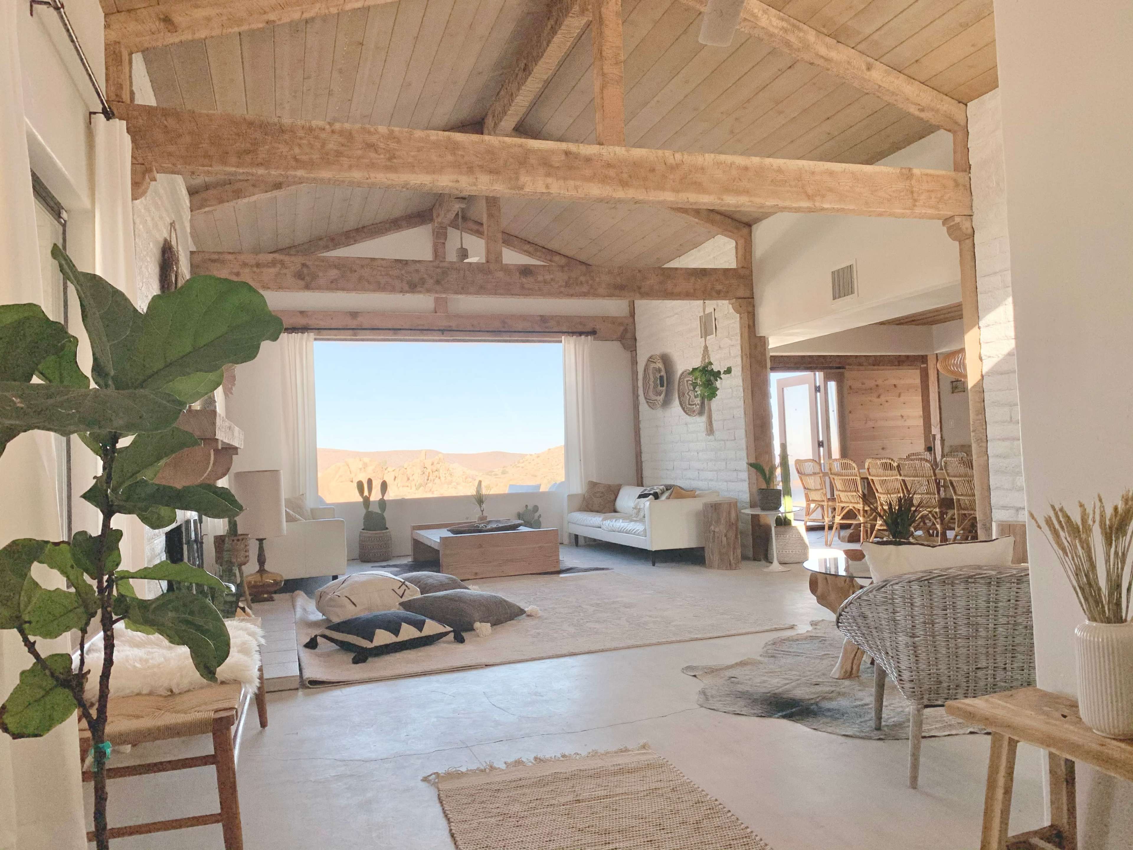 The interior of a spacious, modern living area features wooden beams, a large window with a mountain view, and minimalist furniture.