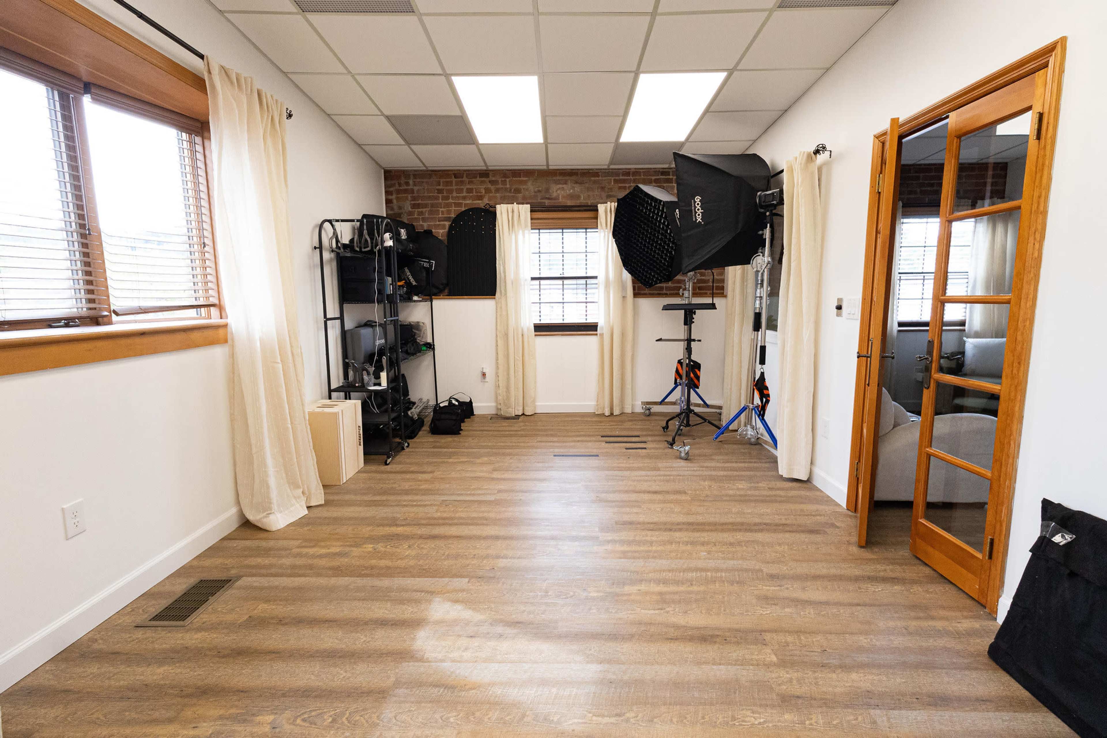 The image shows a bright, empty studio space with wooden floors, large windows, and photography equipment set up in one corner.
