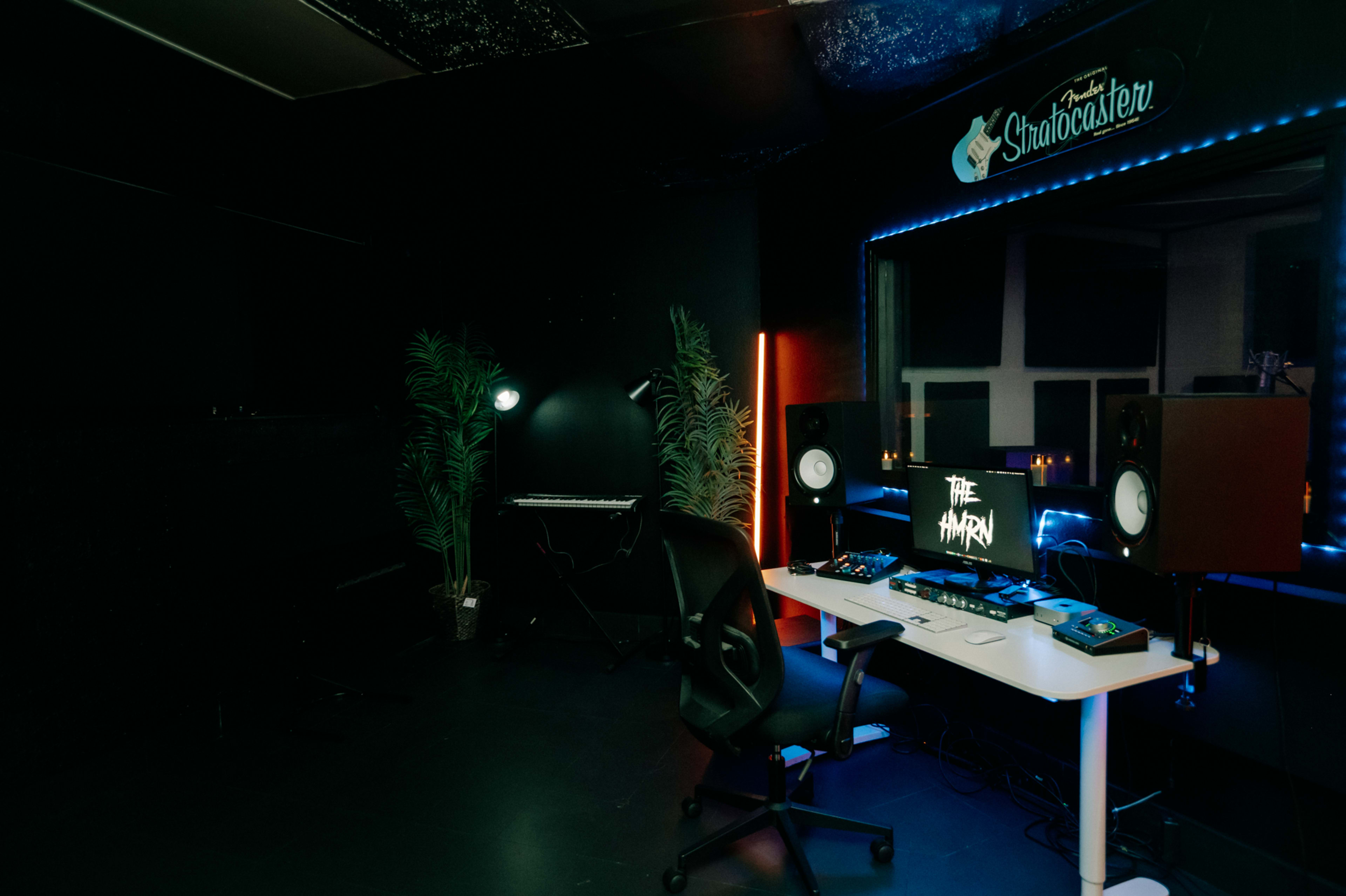 The image shows a music production studio featuring a desk with audio equipment, speakers, and a computer monitor, alongside a keyboard and decorative plants.