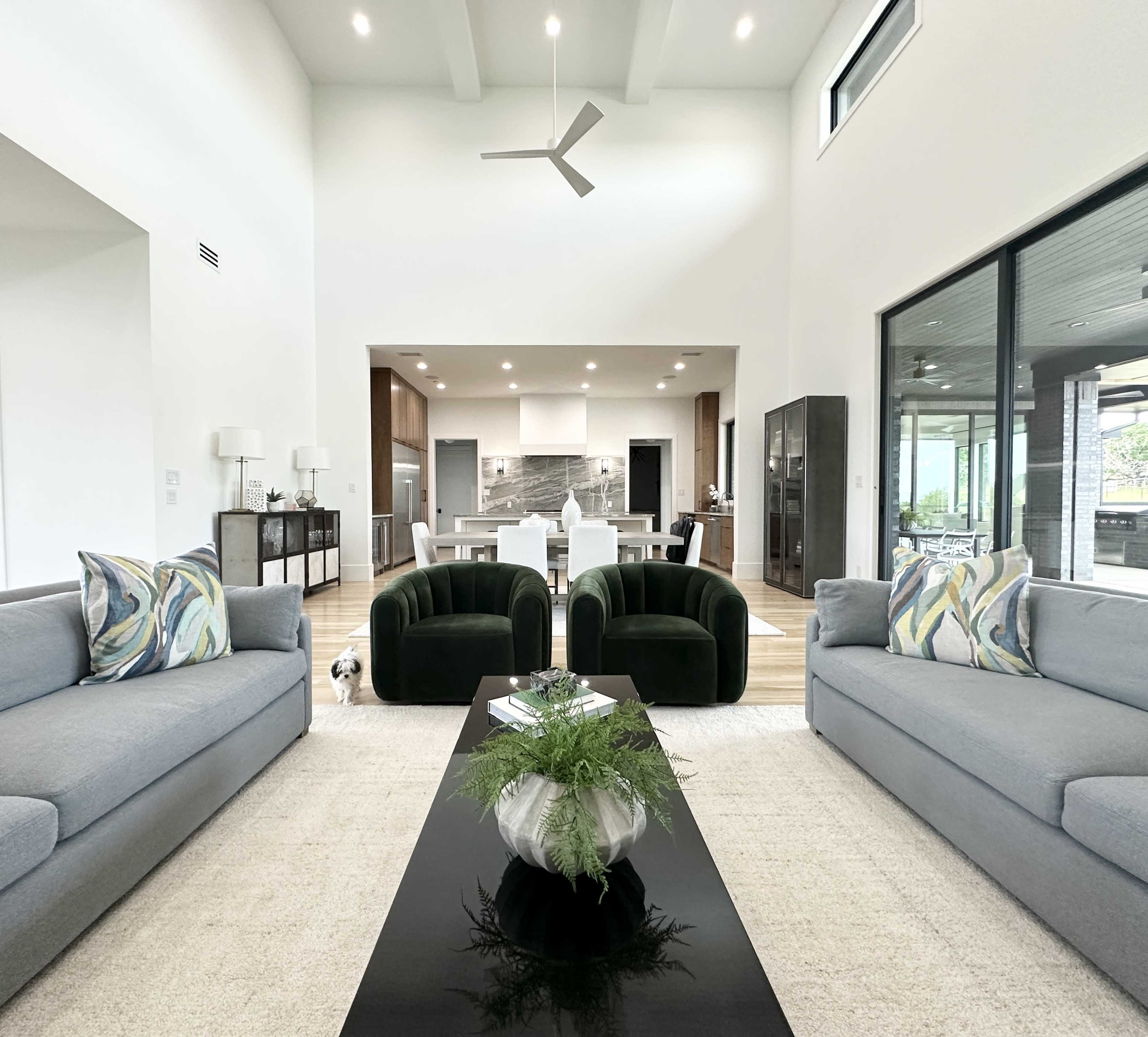 A modern living room features two green armchairs, two gray sofas, a black coffee table, and an open kitchen visible in the background.