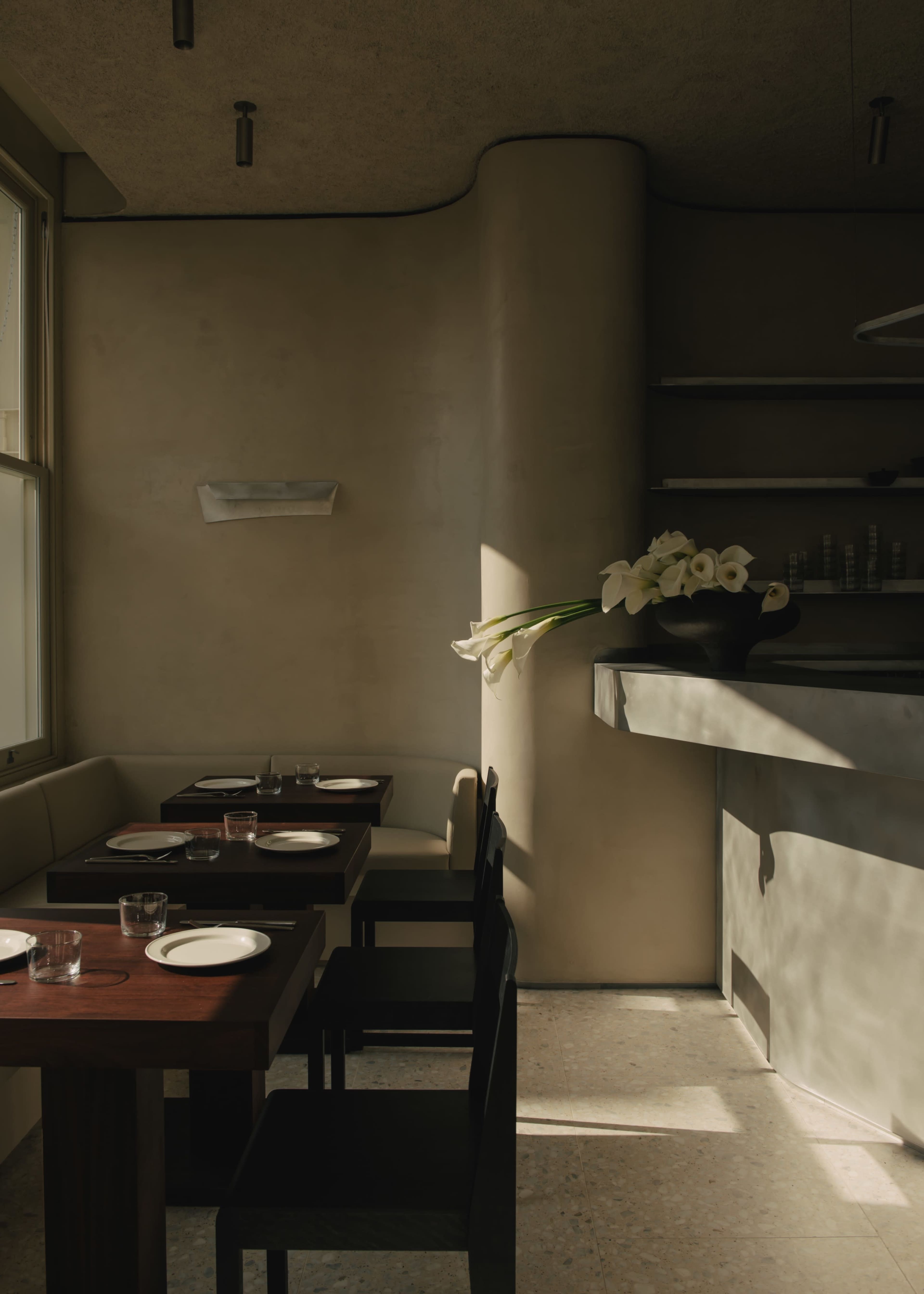 The image shows a minimalist restaurant interior with wooden tables, white plates, and a bar area illuminated by natural light coming through a window.