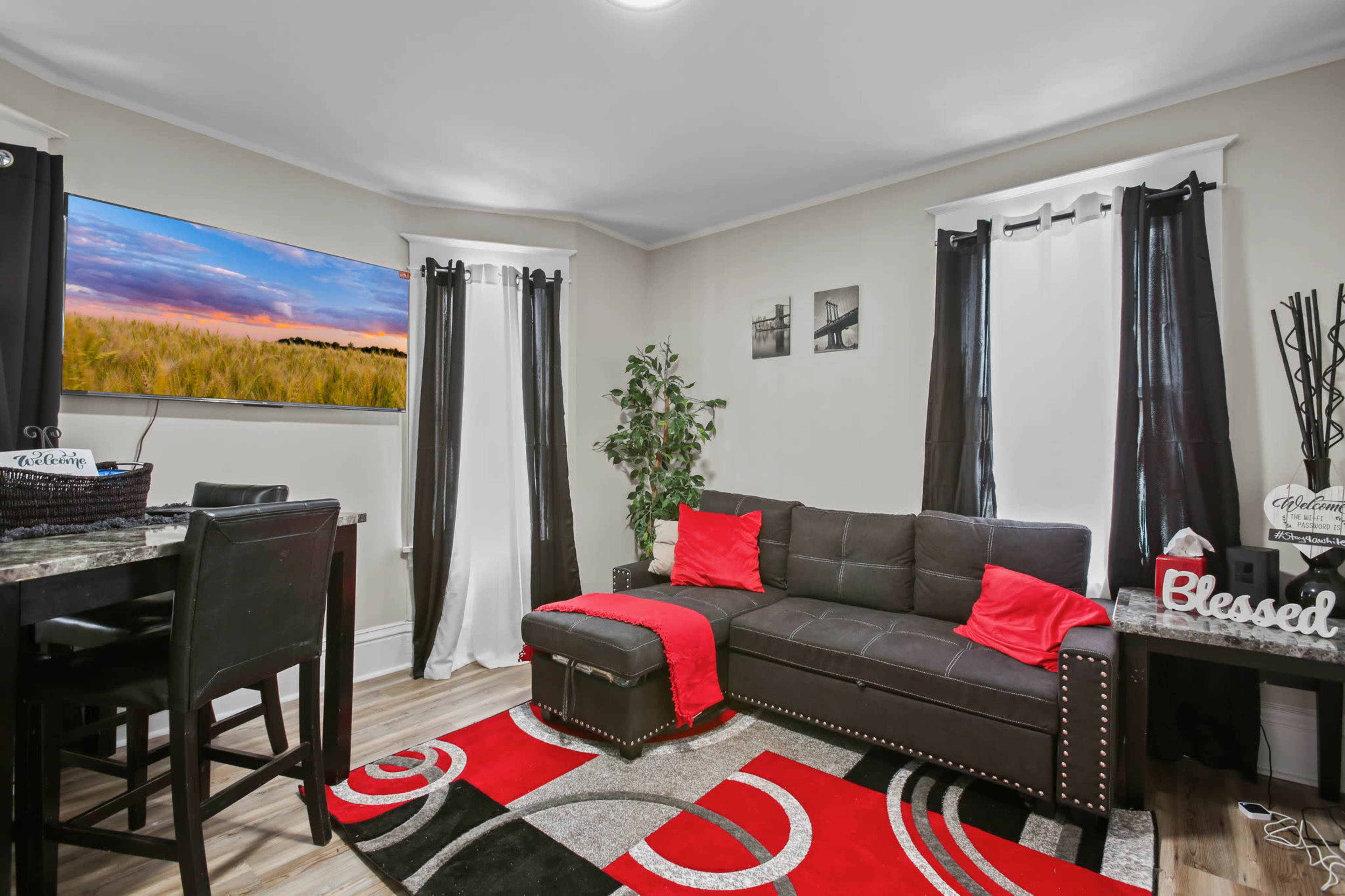 A modern living room features a black sofa with red accents, a dining area with high chairs, and a large TV mounted on the wall displaying a scenic landscape.