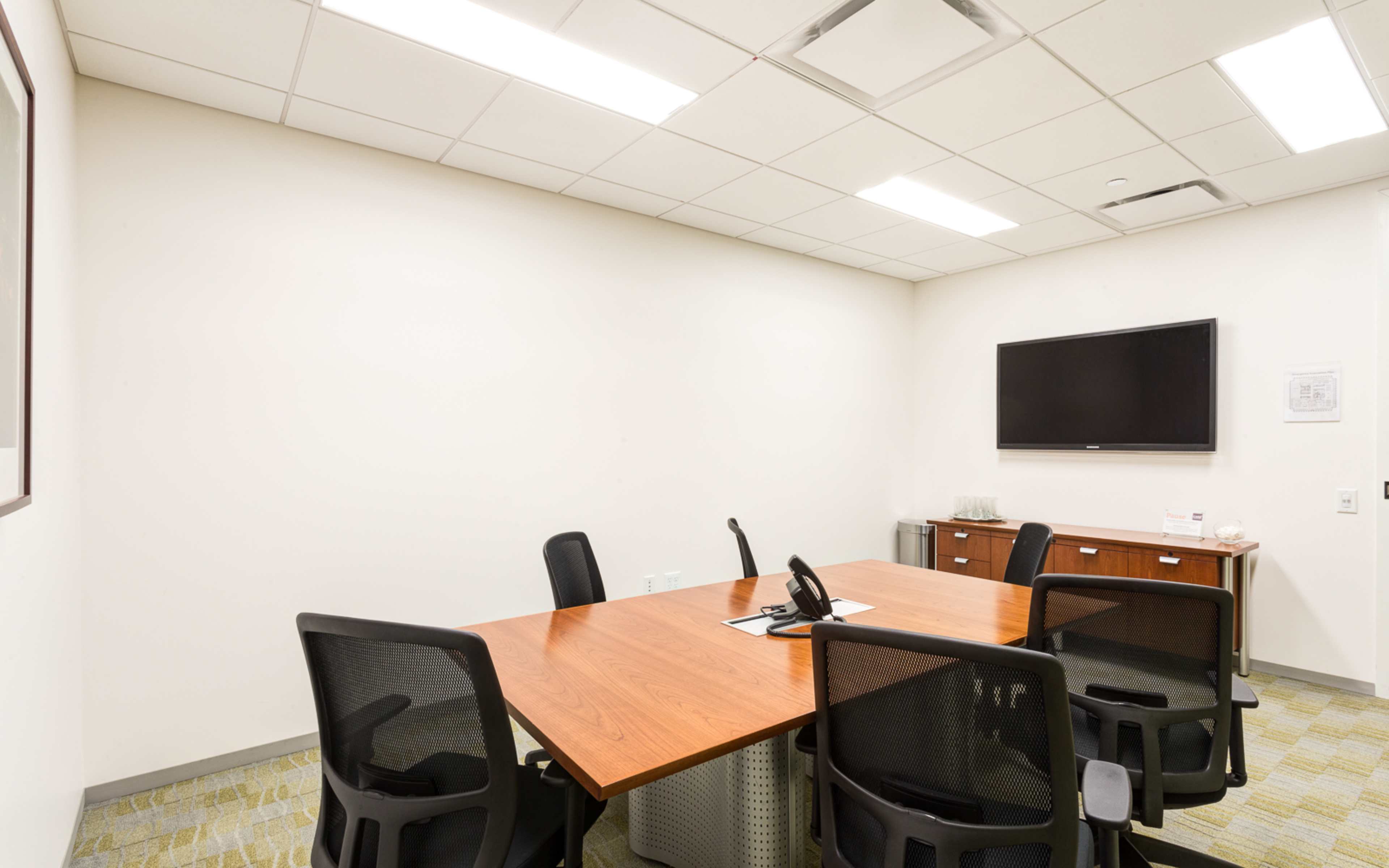 A small conference room features a wooden table surrounded by black chairs, with a wall-mounted television and a cabinet in the background.