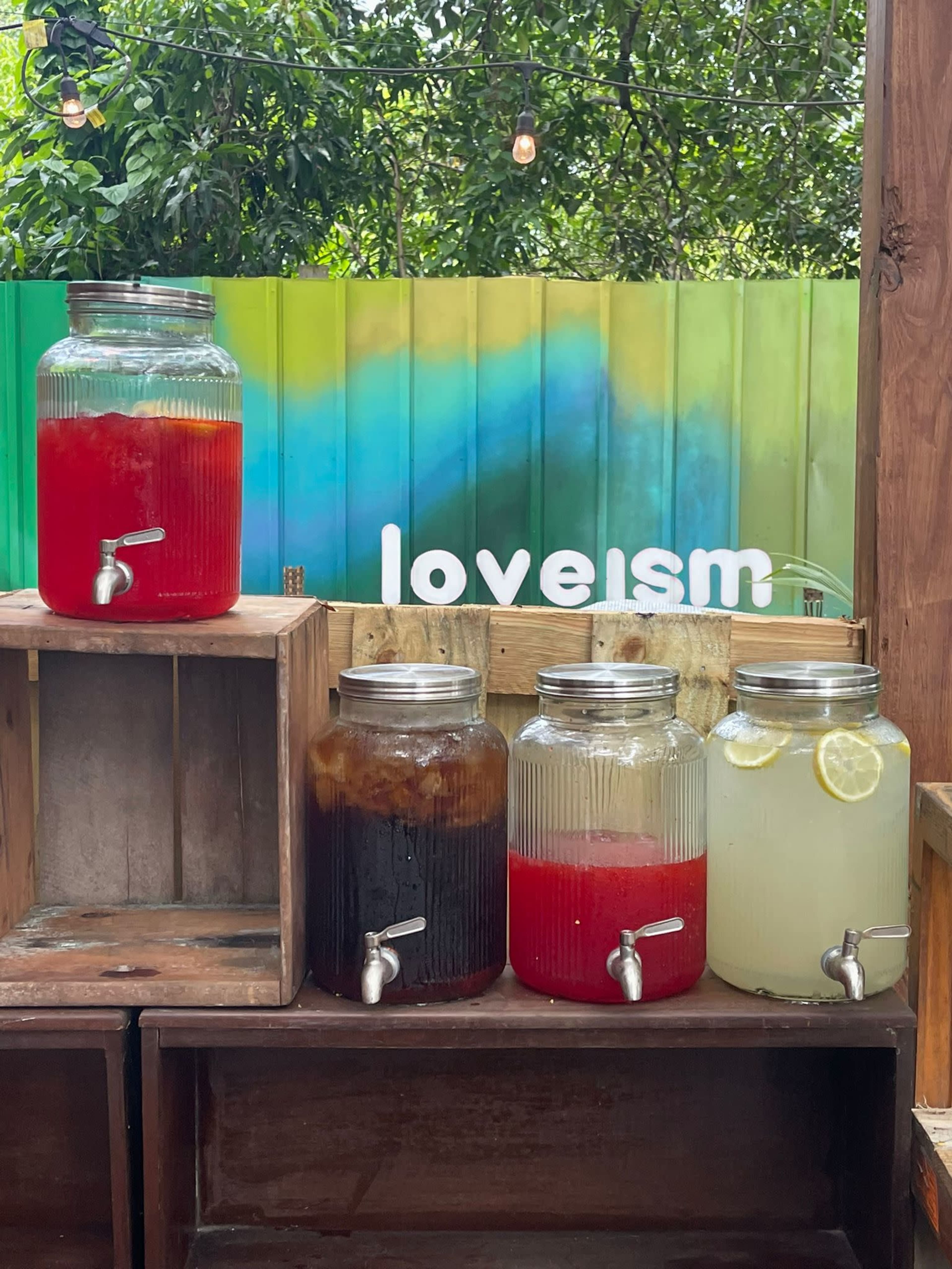 Four glass beverage dispensers filled with colorful drinks are placed on wooden crates against a backdrop of a colorful wall that features the word "loveism."
