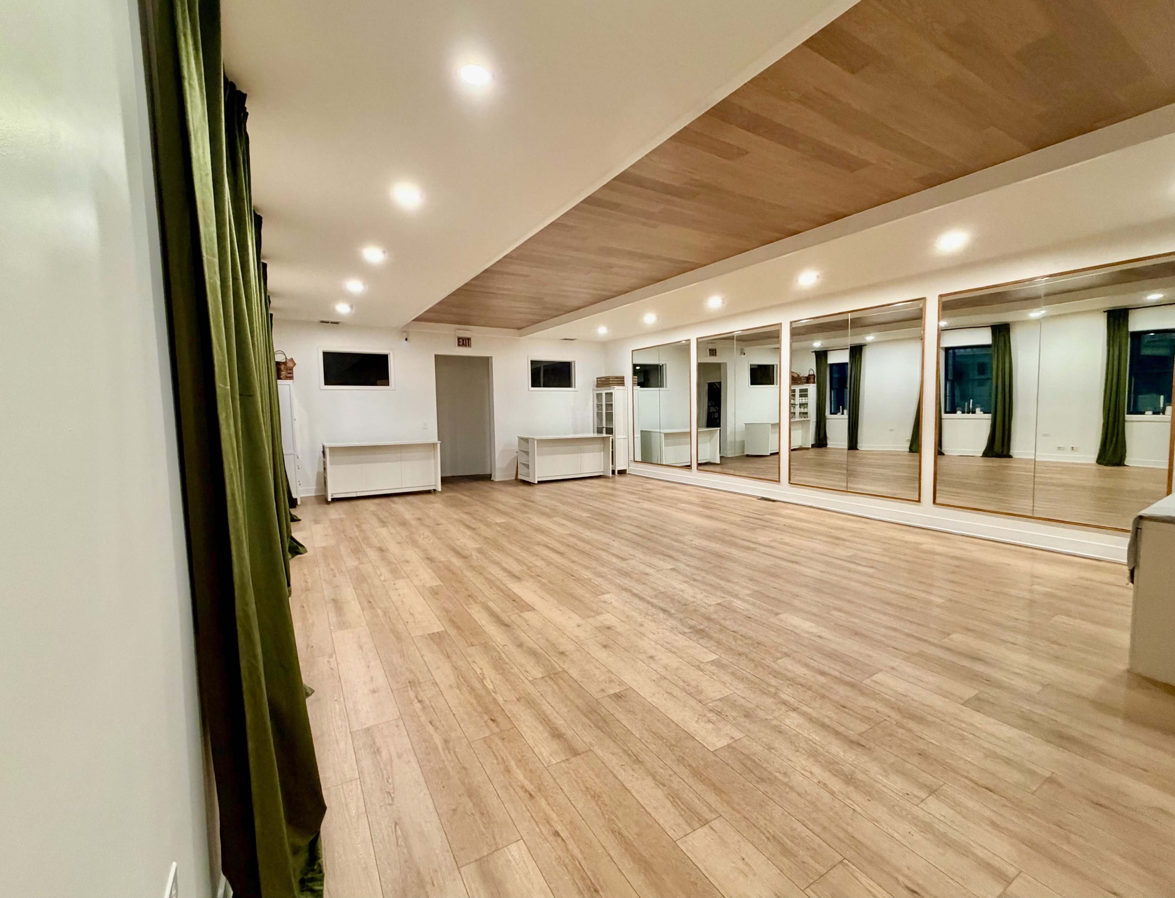 The image shows a spacious room with wooden flooring, large mirrors on one wall, and neutral-colored walls and furnishings.