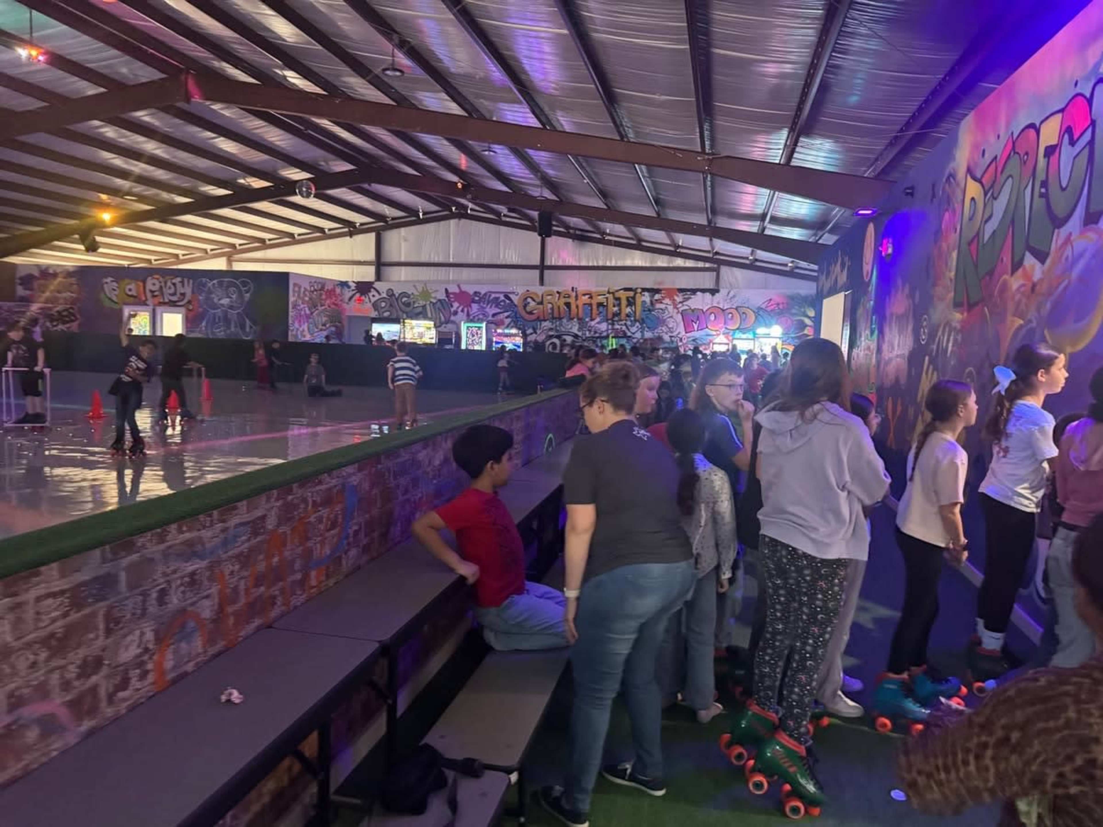 The image shows a roller skating rink with a colorful graffiti wall, where a group of children and adults are gathered near benches, some wearing roller skates.