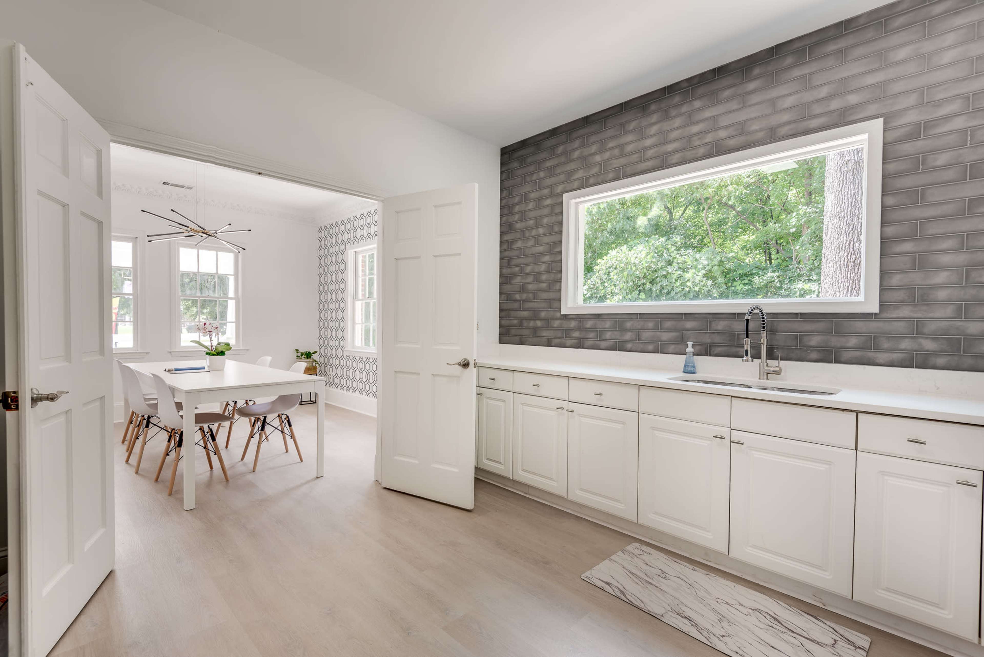 A modern kitchen features white cabinets and a window with a view of greenery, adjacent to a dining area with a large table and stylish chairs.