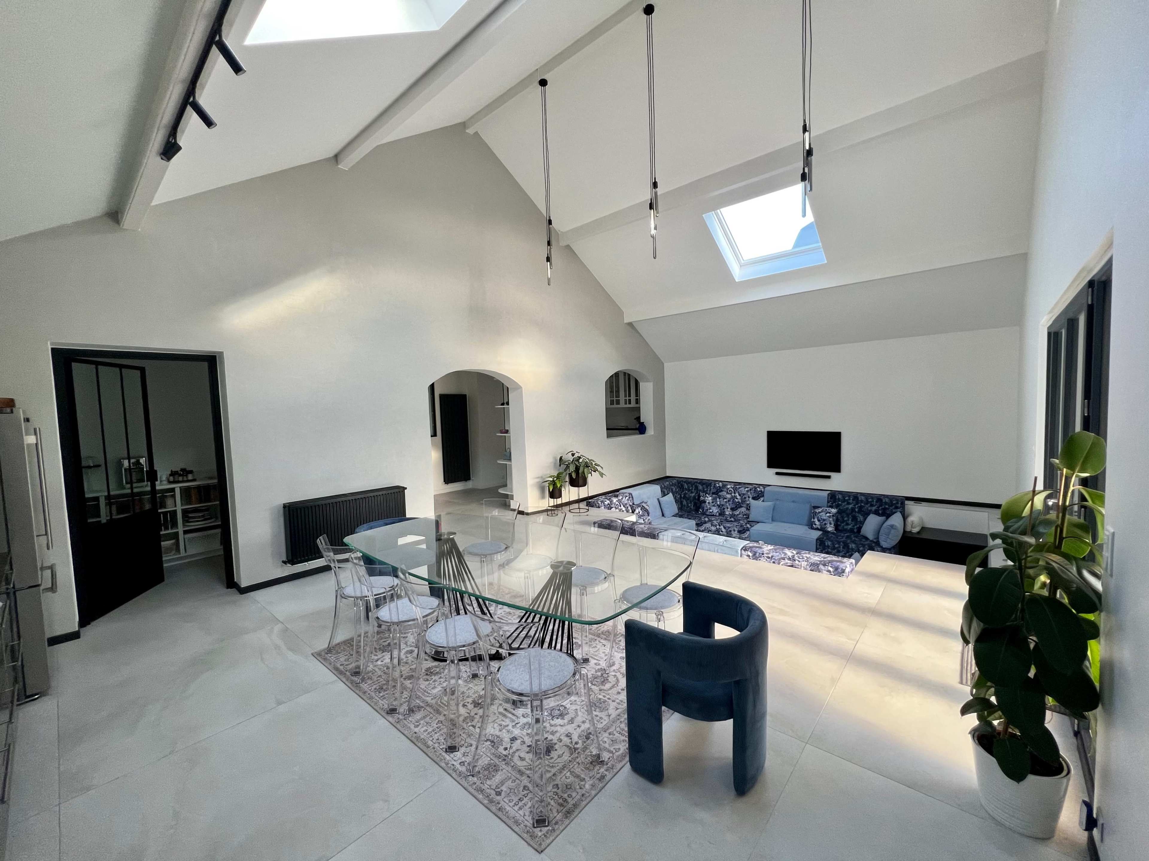 A spacious dining area features a glass table surrounded by transparent chairs, with a skylight illuminating the room and a modern sectional sofa in the background.