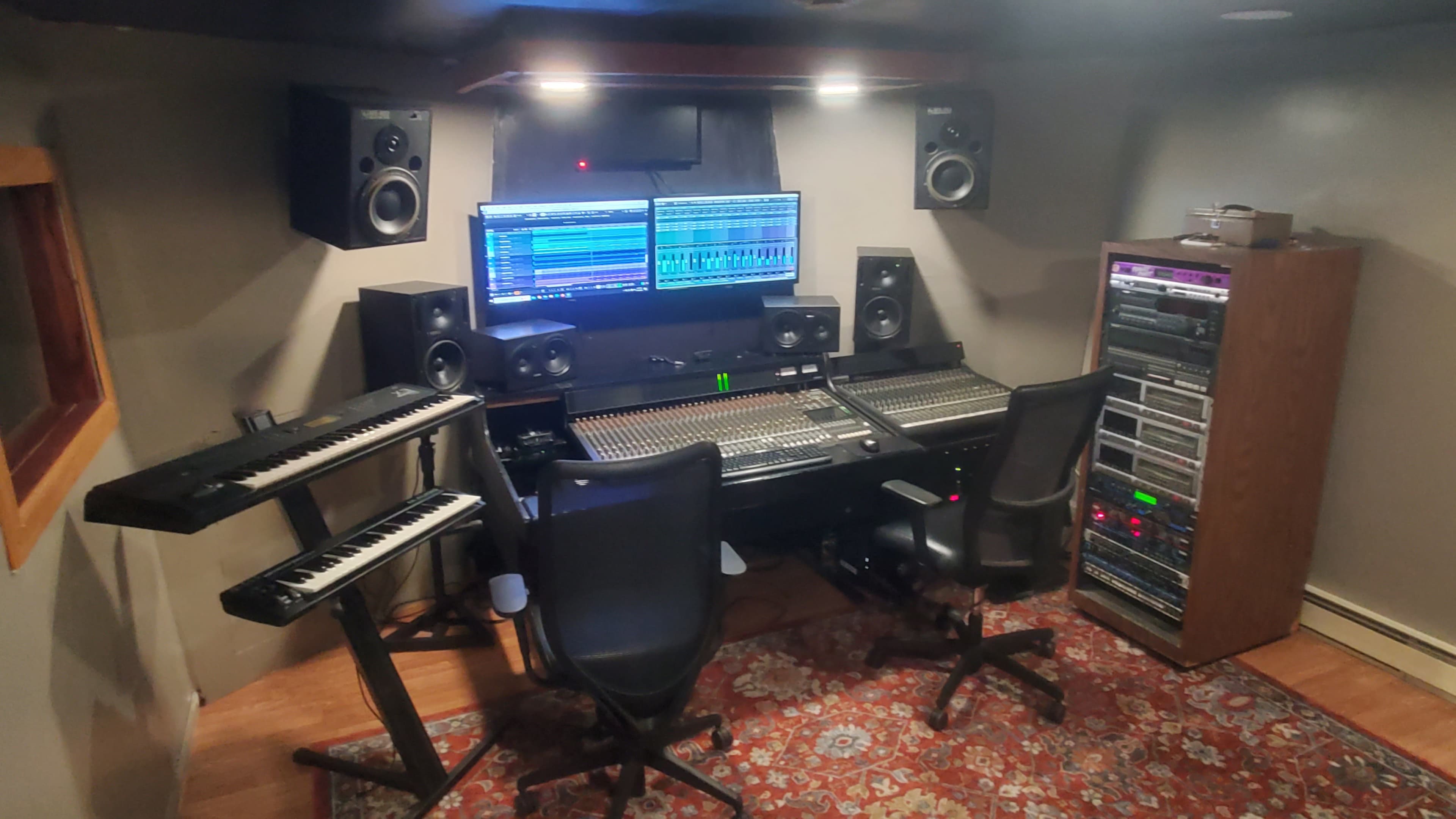 A music recording studio featuring mixing equipment, two computer monitors, audio speakers, and synthesizers arranged around a central mixing console.