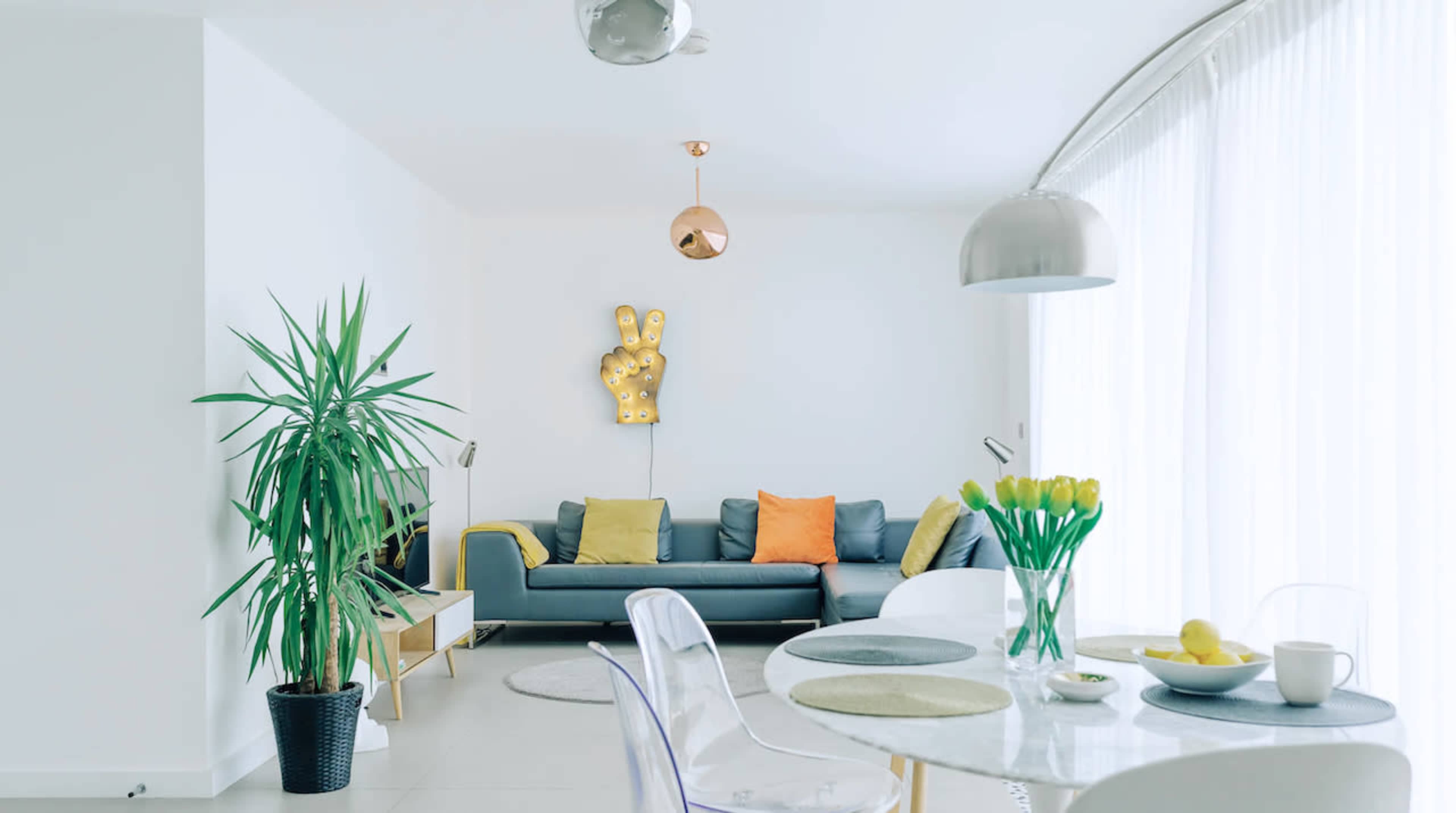 A modern living space features a grey sofa with colorful cushions, a dining table set with tulips and fruit, and a large plant in the corner.