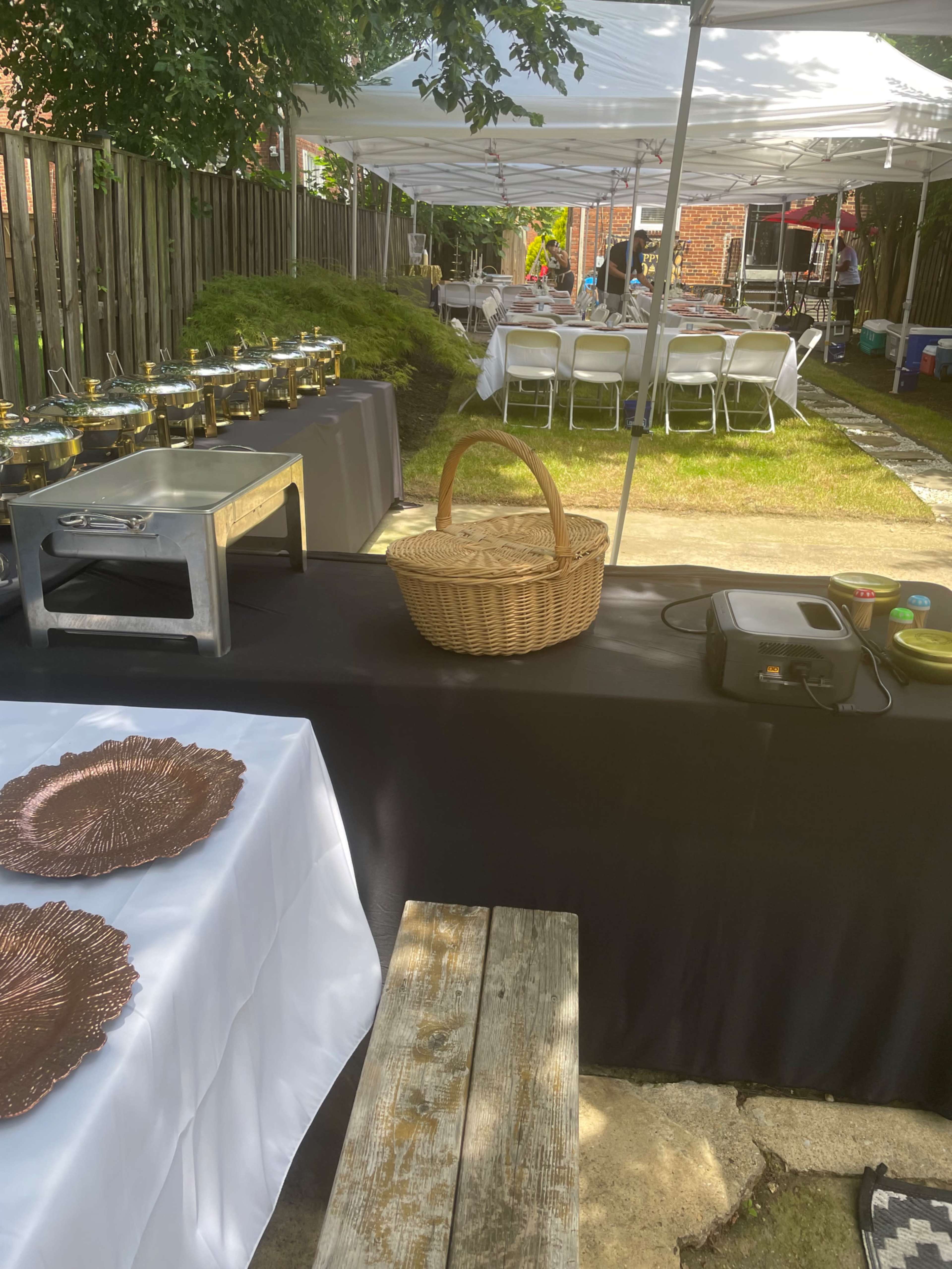 A backyard setting features a table with food service items, a wicker basket, and folding chairs under tents in the background.