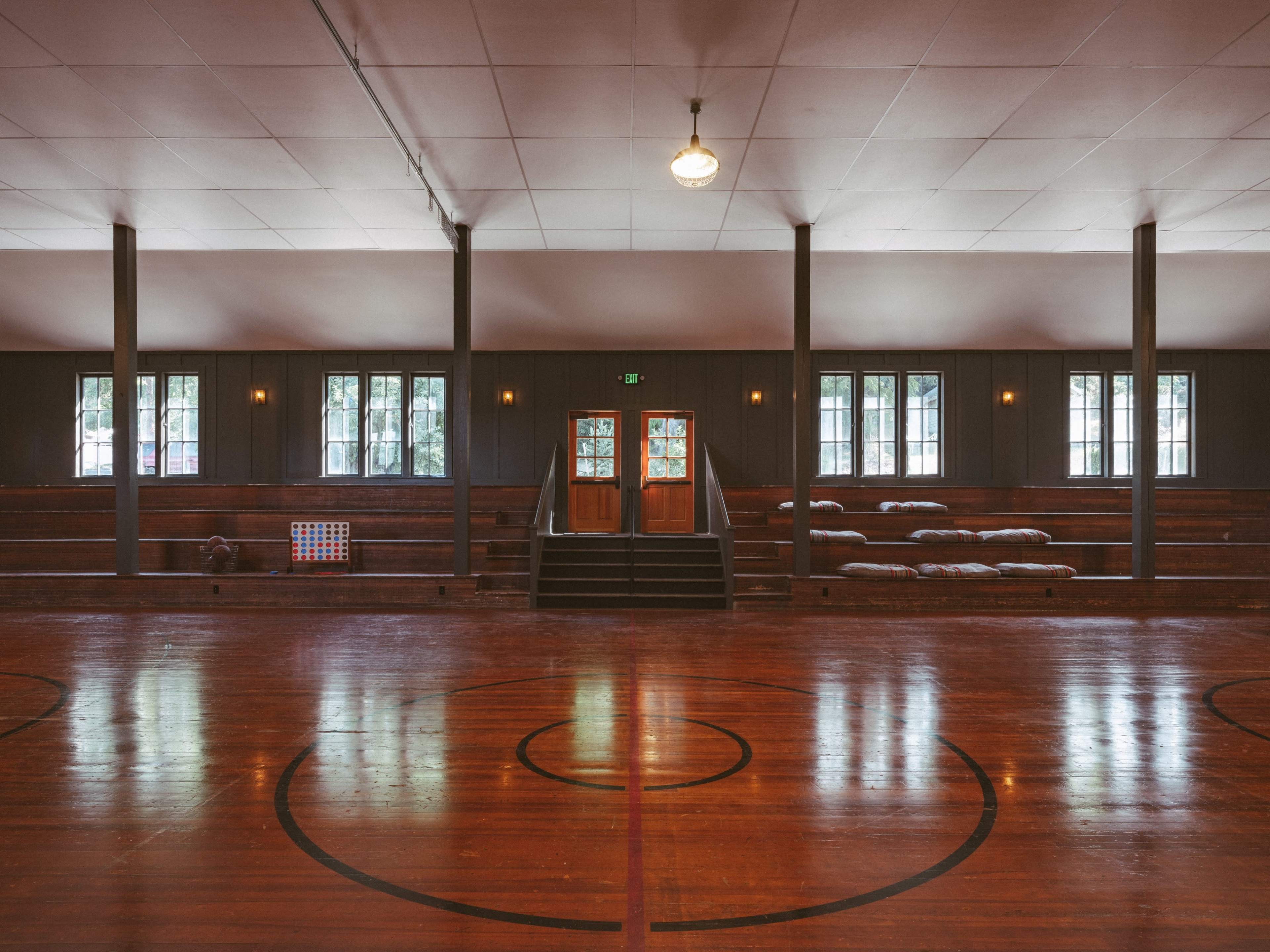 The image shows an empty wooden gymnasium with polished floors, large windows, and bleachers along the walls.