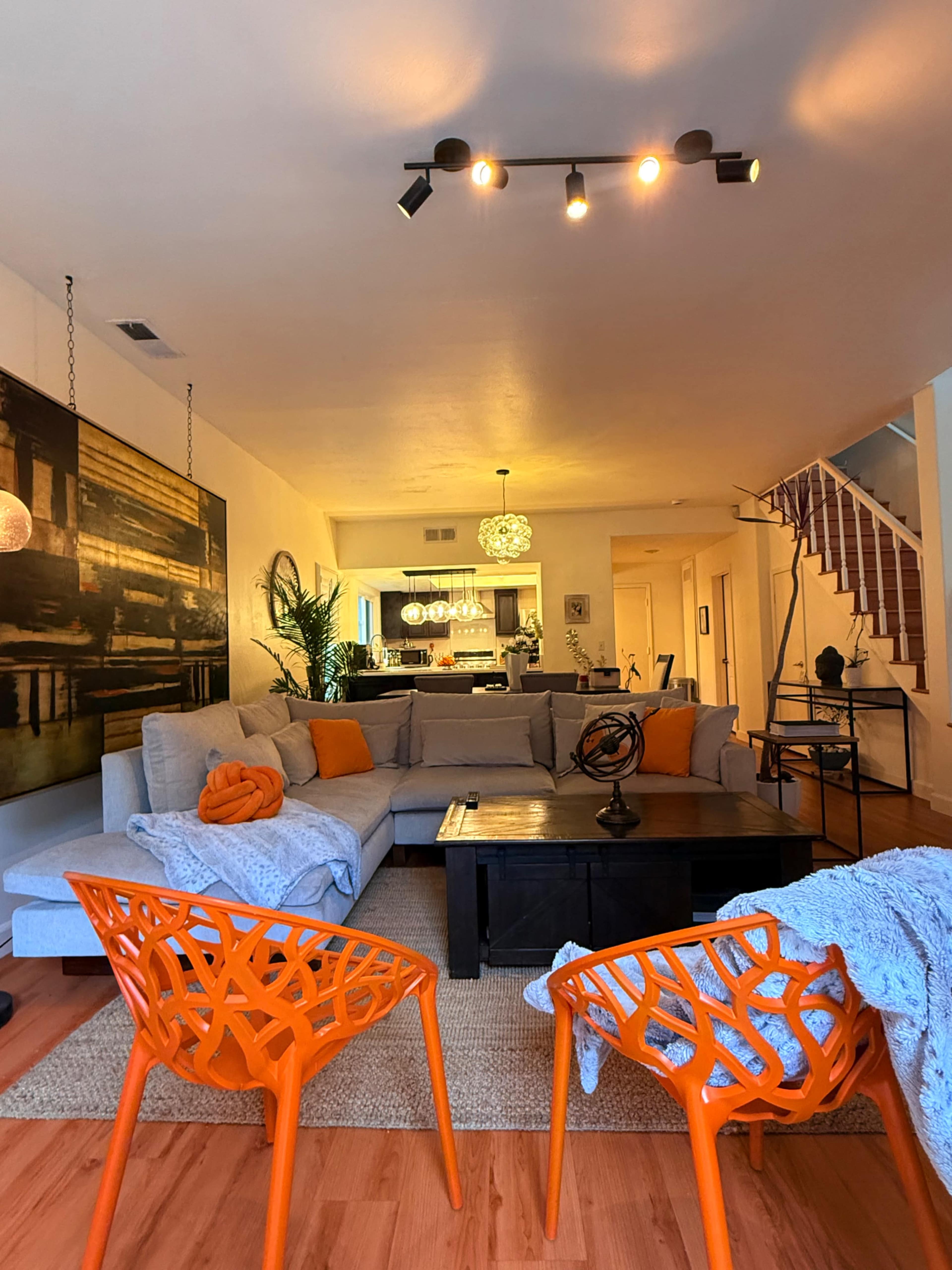 The living room features a sectional sofa with colorful throw pillows, orange chairs, and a coffee table, all arranged around a woven area rug, leading to a kitchen visible in the background.