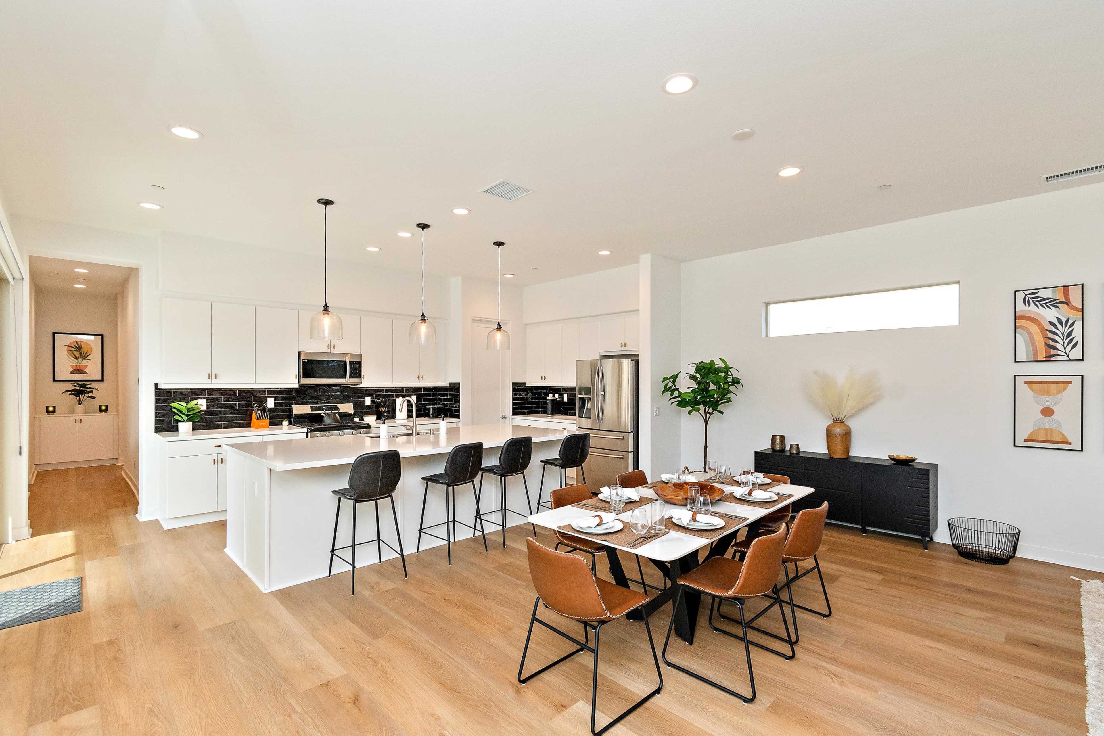 A modern kitchen and dining area features white cabinetry, black tile backsplash, and a large dining table set with dishes, surrounded by stylish chairs.