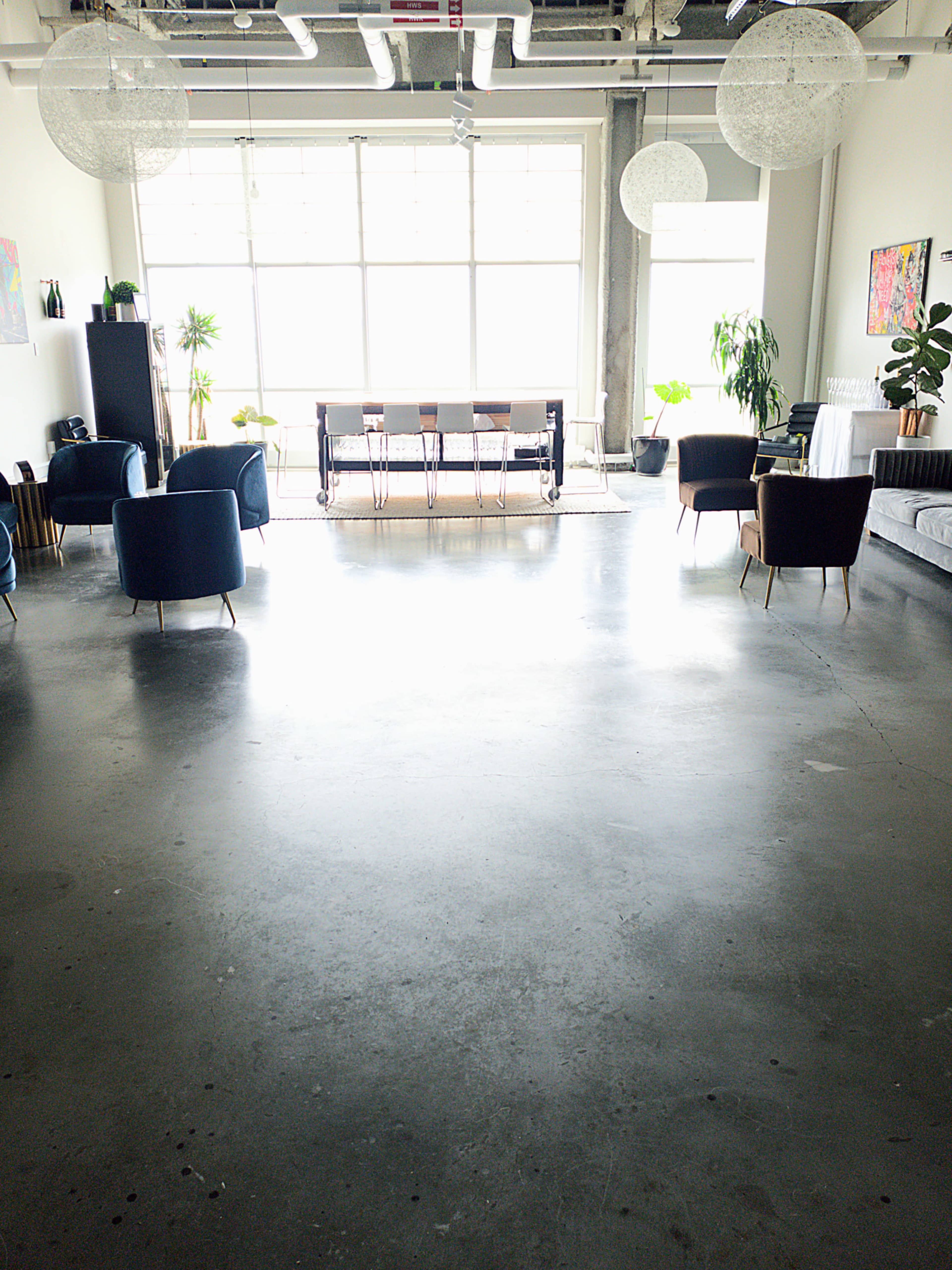The image shows a spacious, modern interior with a concrete floor, several contemporary chairs, a dining table, and large windows allowing natural light.