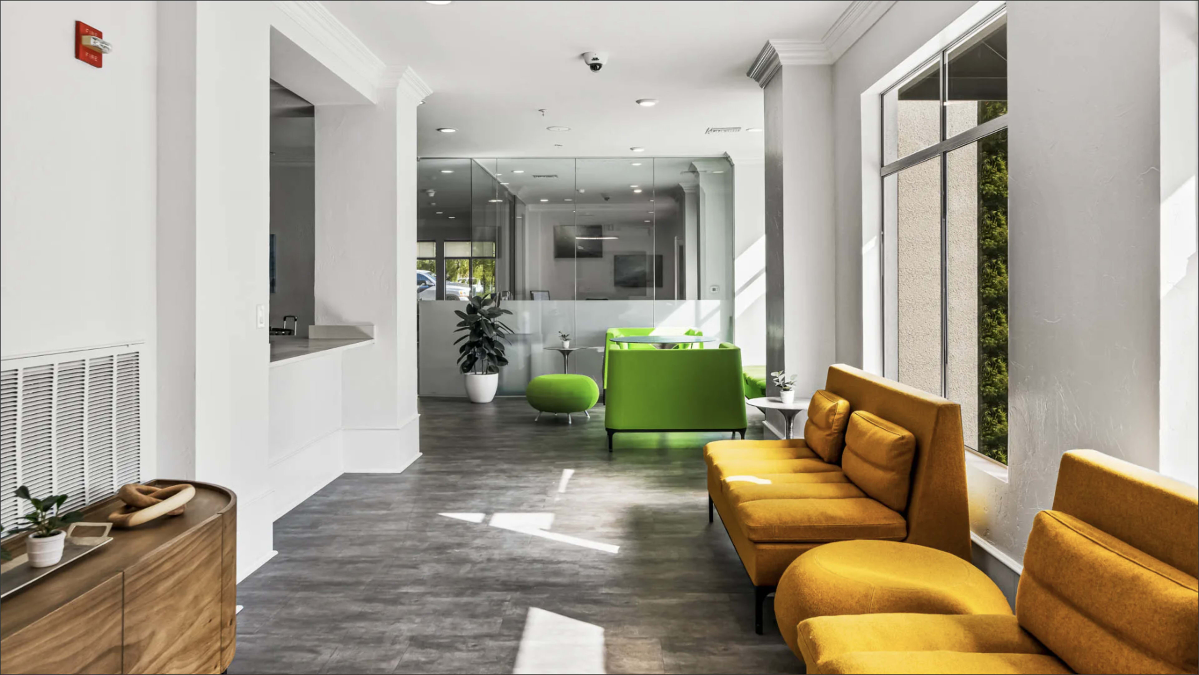 A bright, modern waiting area features yellow and green seating, large windows letting in natural light, and contemporary decor.