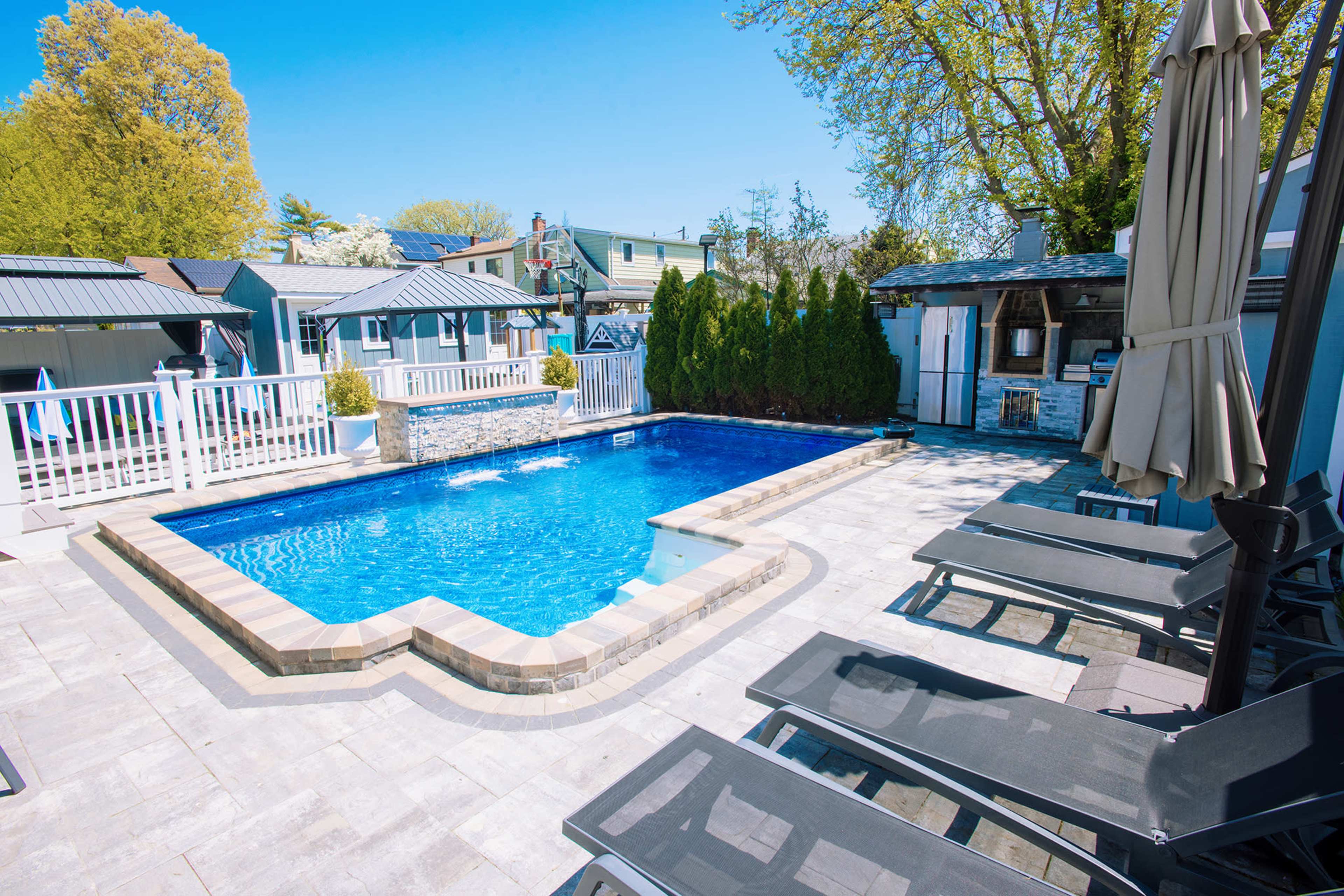 The image shows a rectangular swimming pool surrounded by a stone patio and lounge chairs, with nearby greenery and structures in the background.