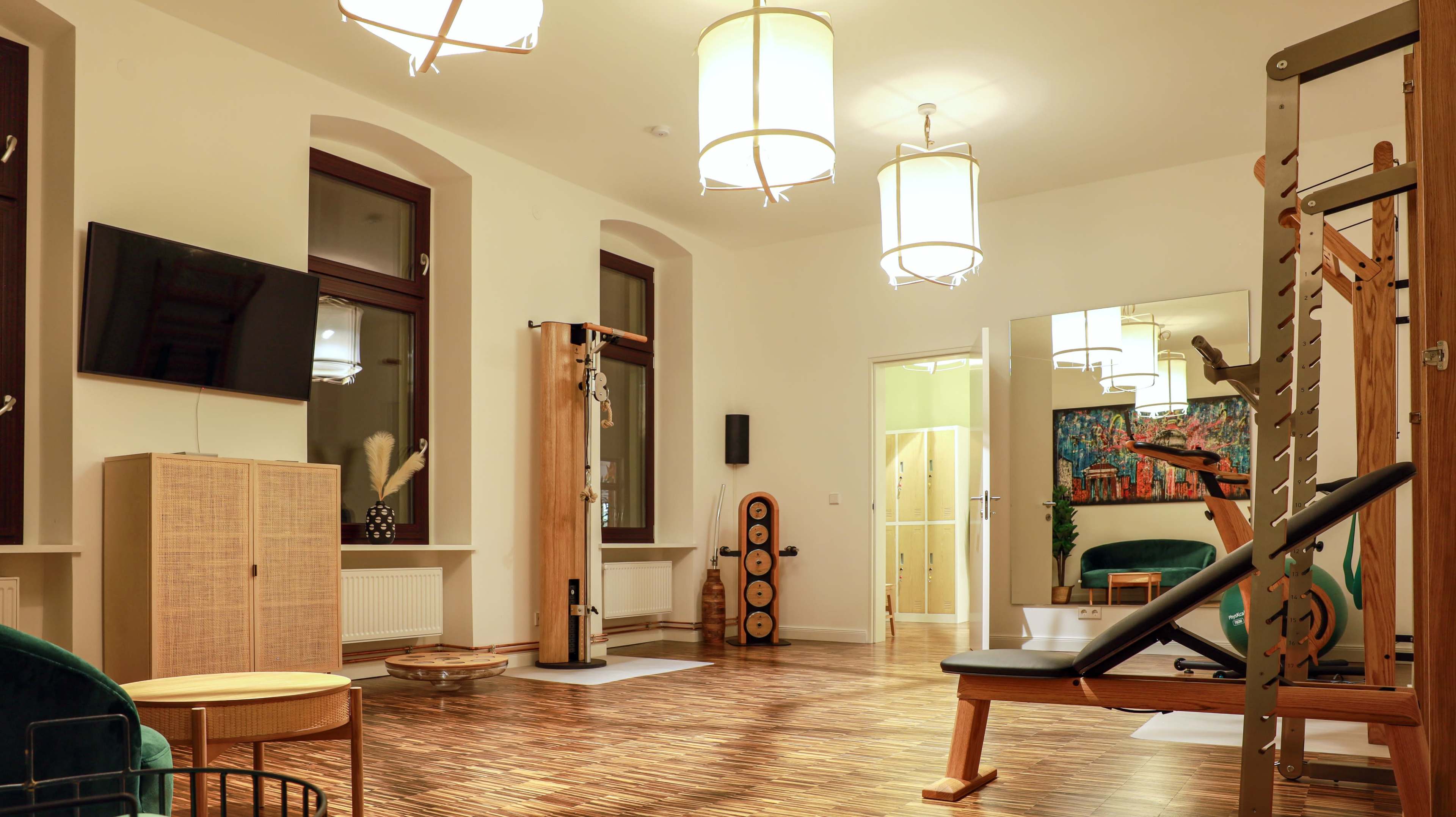 The image shows a spacious indoor fitness studio with wooden floors, multiple light fixtures, exercise equipment, and a large mirror.