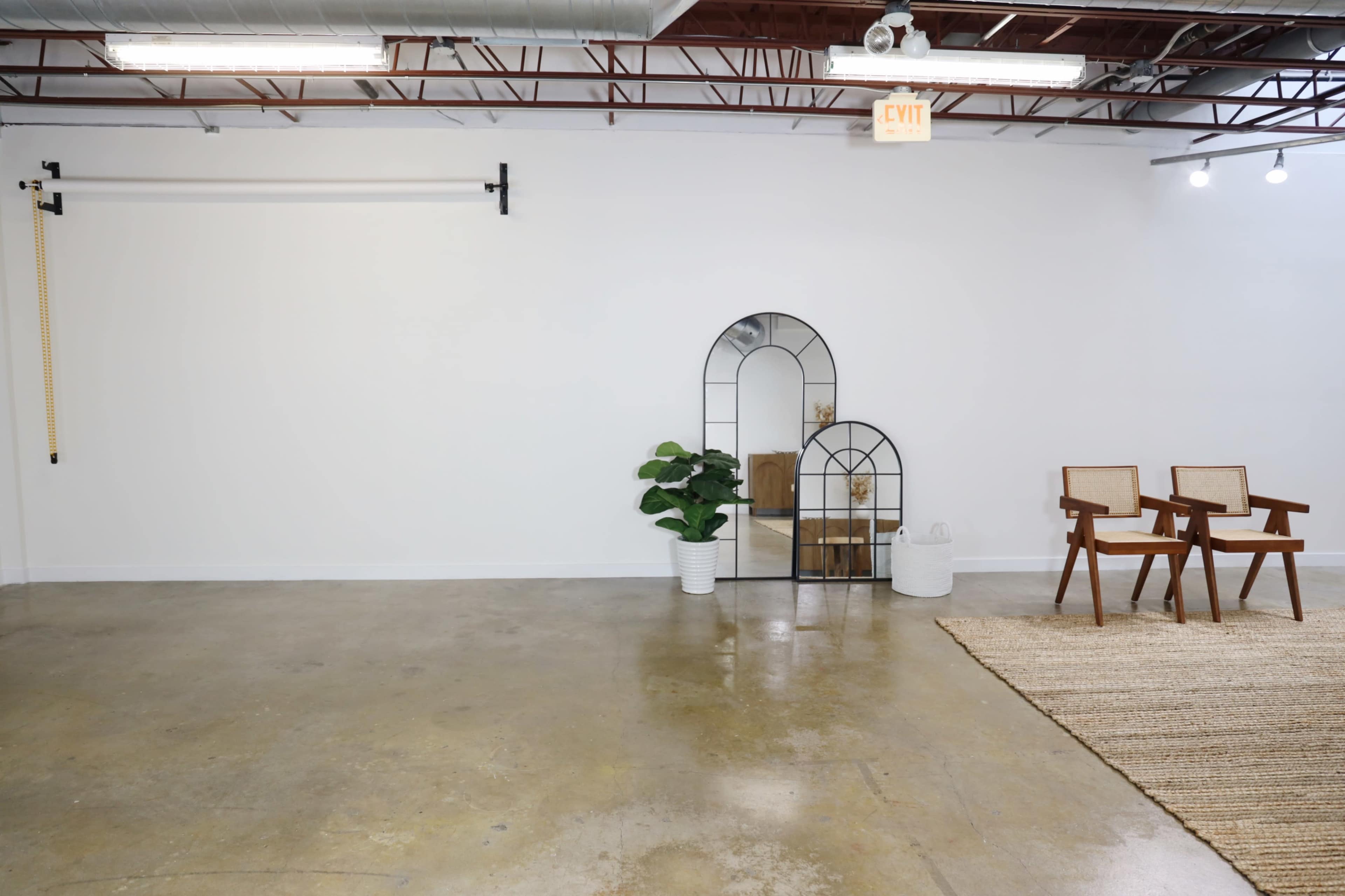 A minimalistic interior space with a concrete floor, a white wall, a decorative archway with a plant, two wooden chairs, and a rug on the floor.