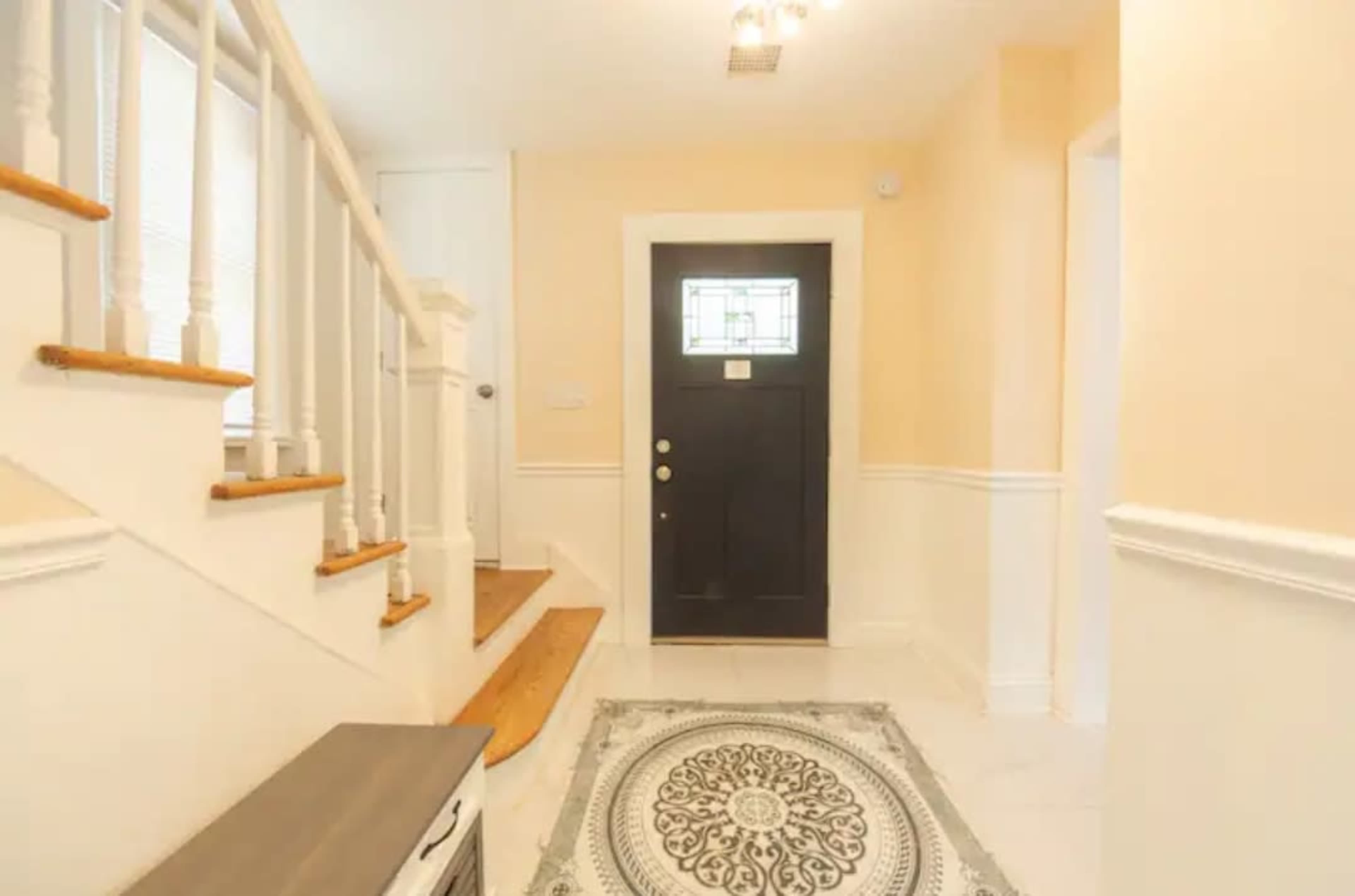 A foyer with a staircase on the left, a black front door at the center, and a patterned rug on the tiled floor.