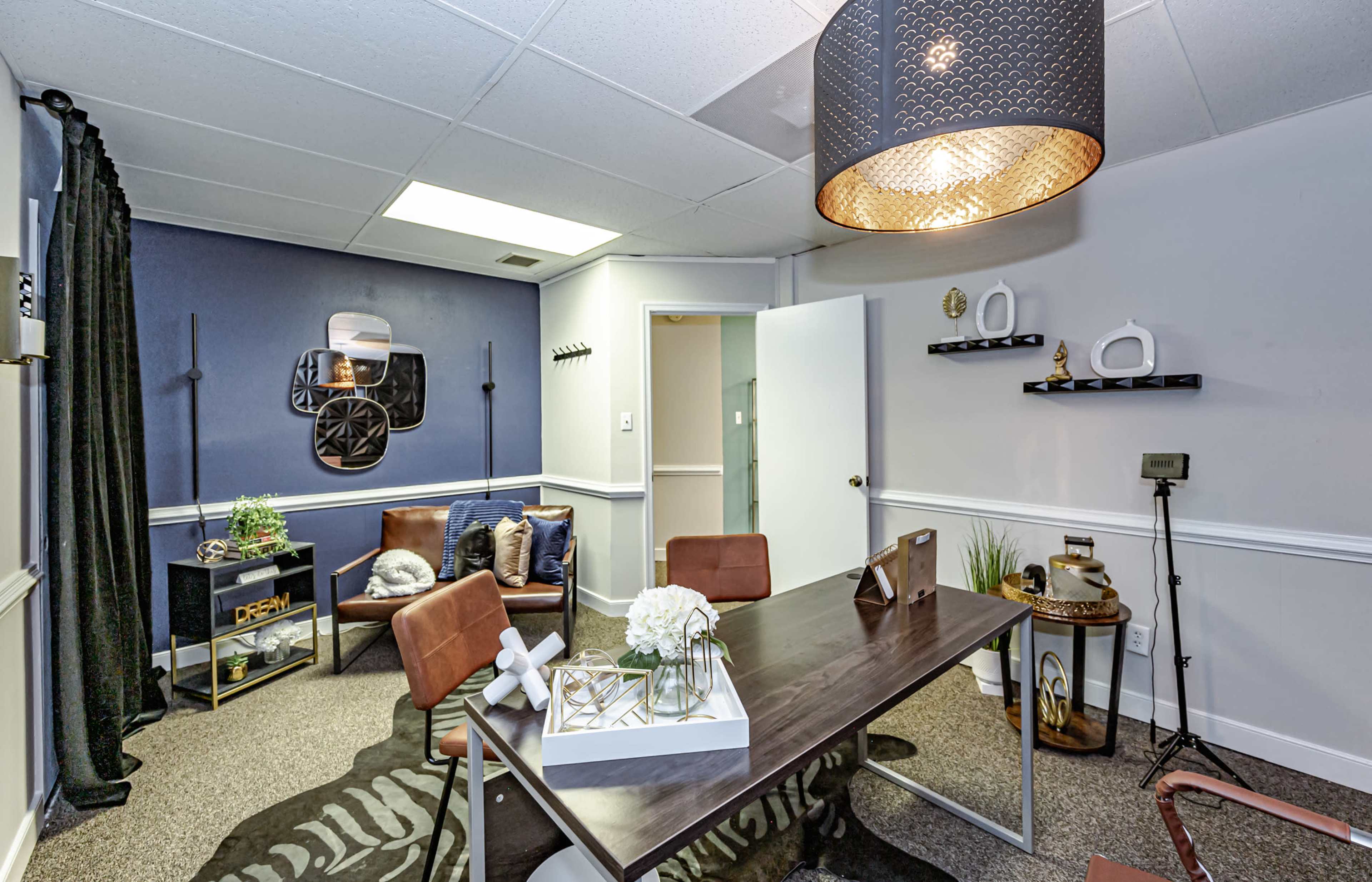 A modern office space features a table, chairs, decorative items, and a large overhead light, with dark blue walls and carpeting.