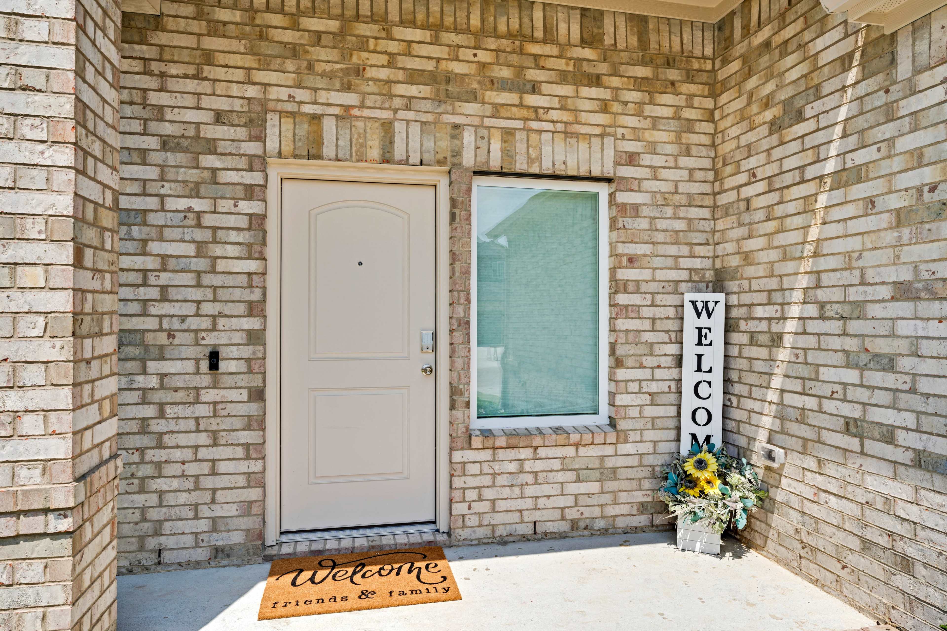 A front porch with a light-colored door, a welcome mat, and a decorative sign surrounded by flowers.